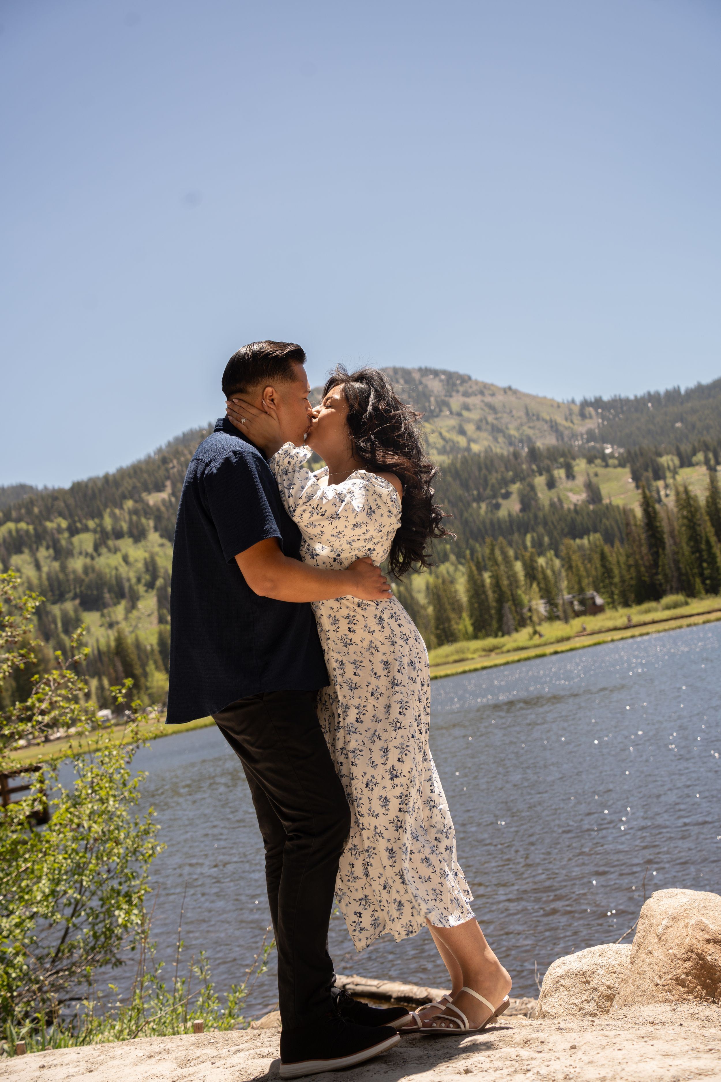 Couple sharing a romantic kiss by silver lake Utah on a sunny day, surrounded by lush green trees and hills.