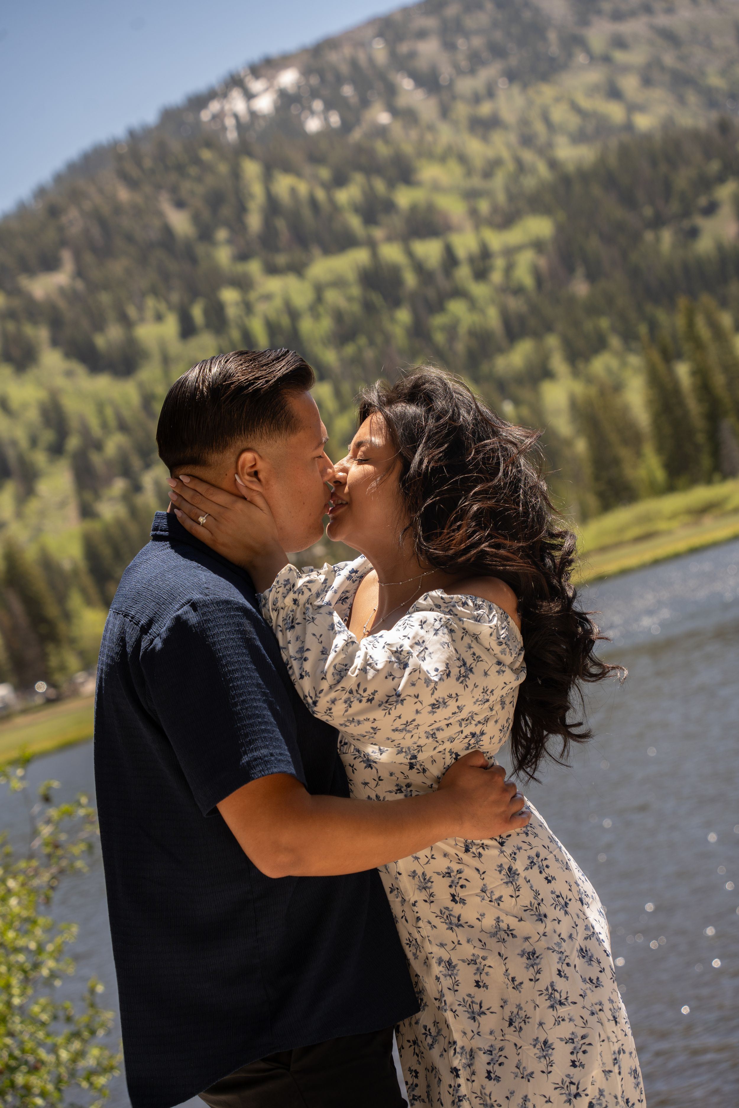 A couple shares a romantic kiss by Silver lake, with lush green trees and a scenic peak in the background.
