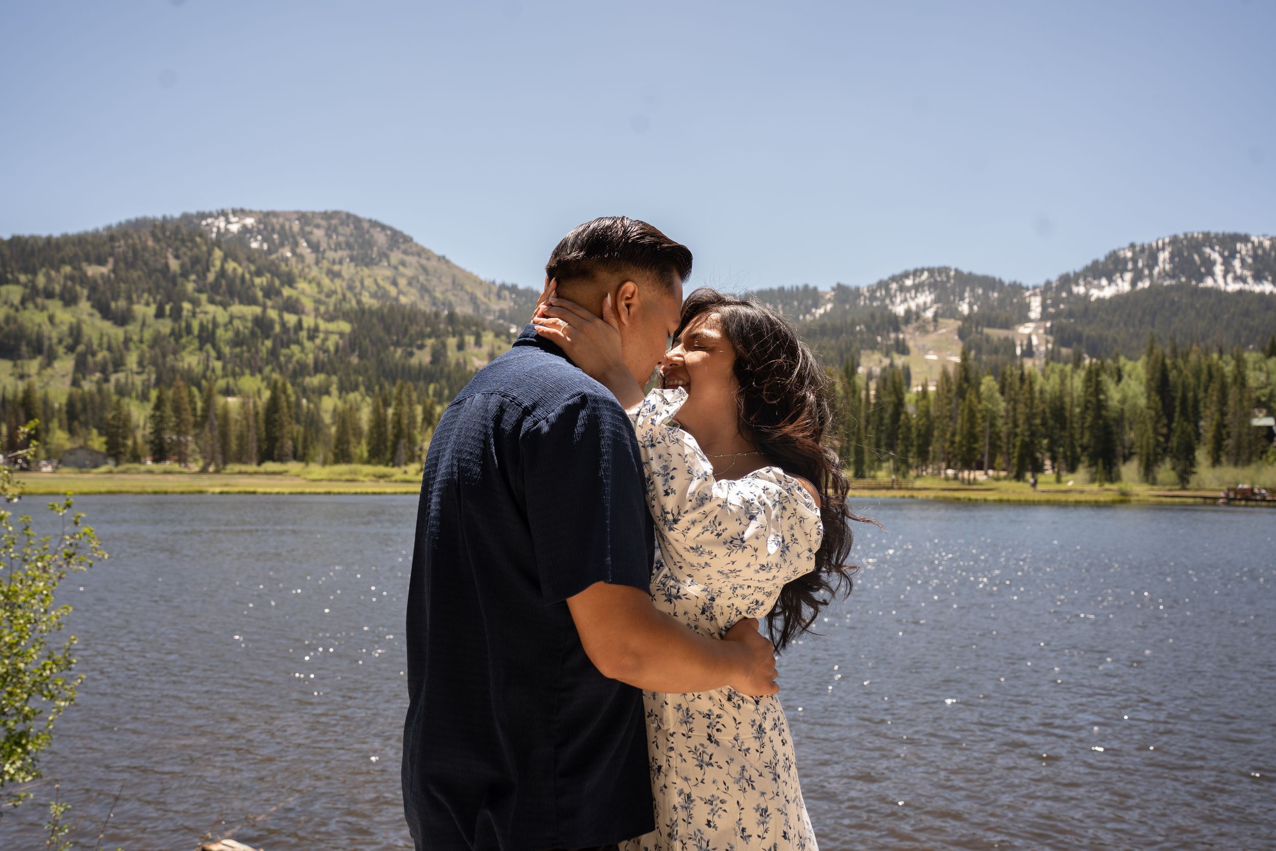 Couple sharing a romantic moment by a mountain lake surrounded by pine trees and snow-capped peaks on a sunny day.