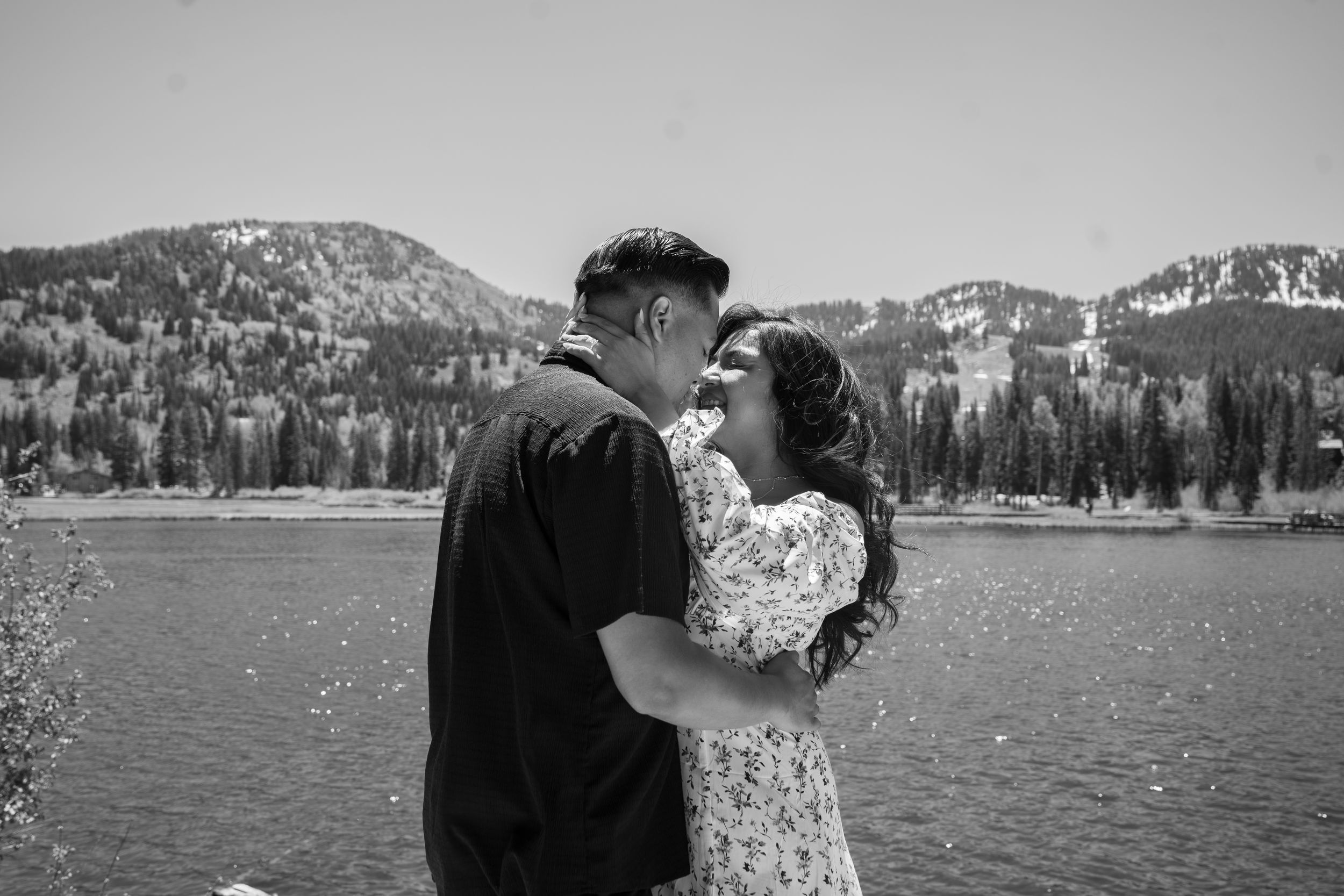 Couple sharing a romantic moment by silver lake with snow-capped peaks and pine trees in black and white.
