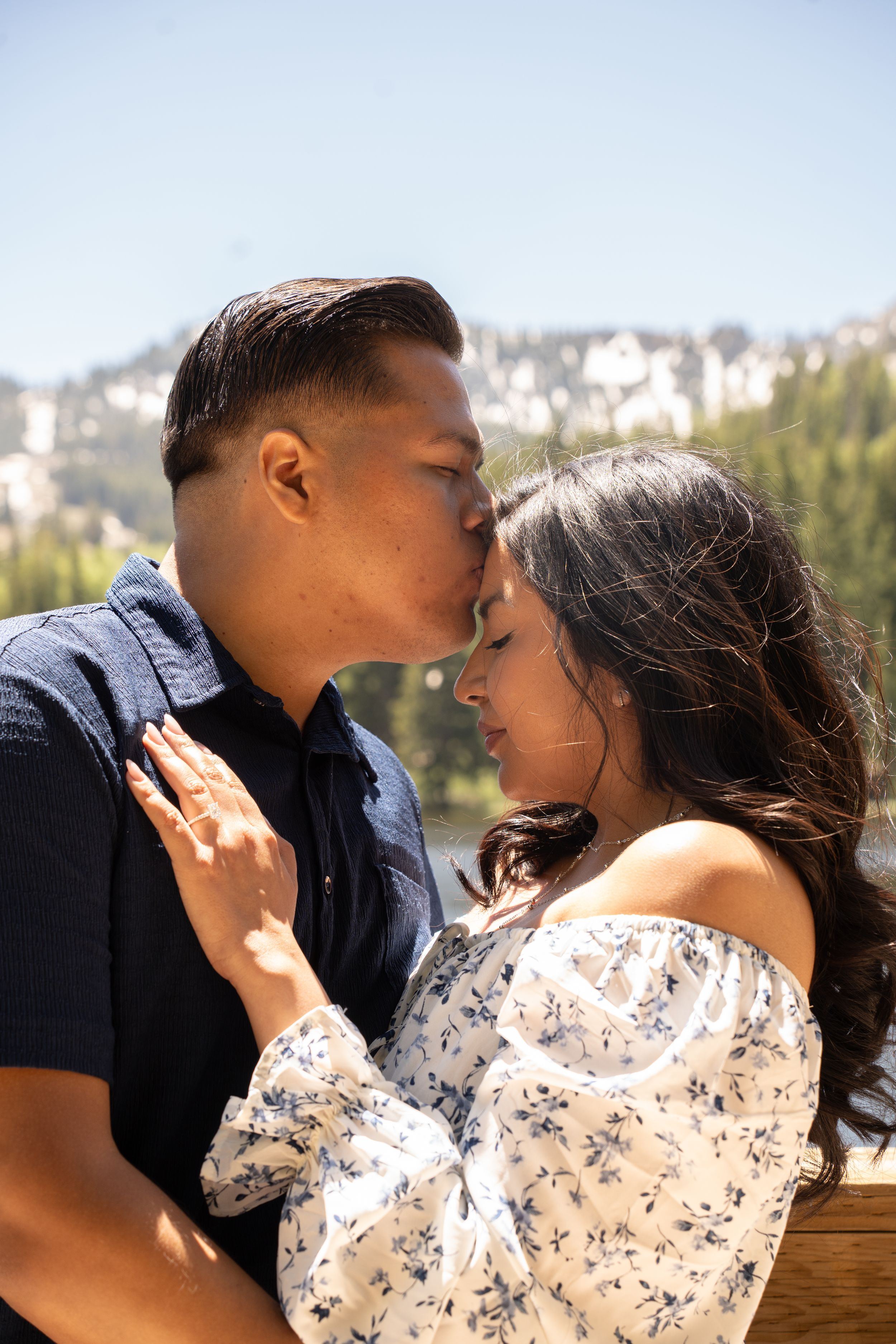 A couple shares a tender forehead kiss at silver lake, the woman wearing a floral off-shoulder top and engagement ring.