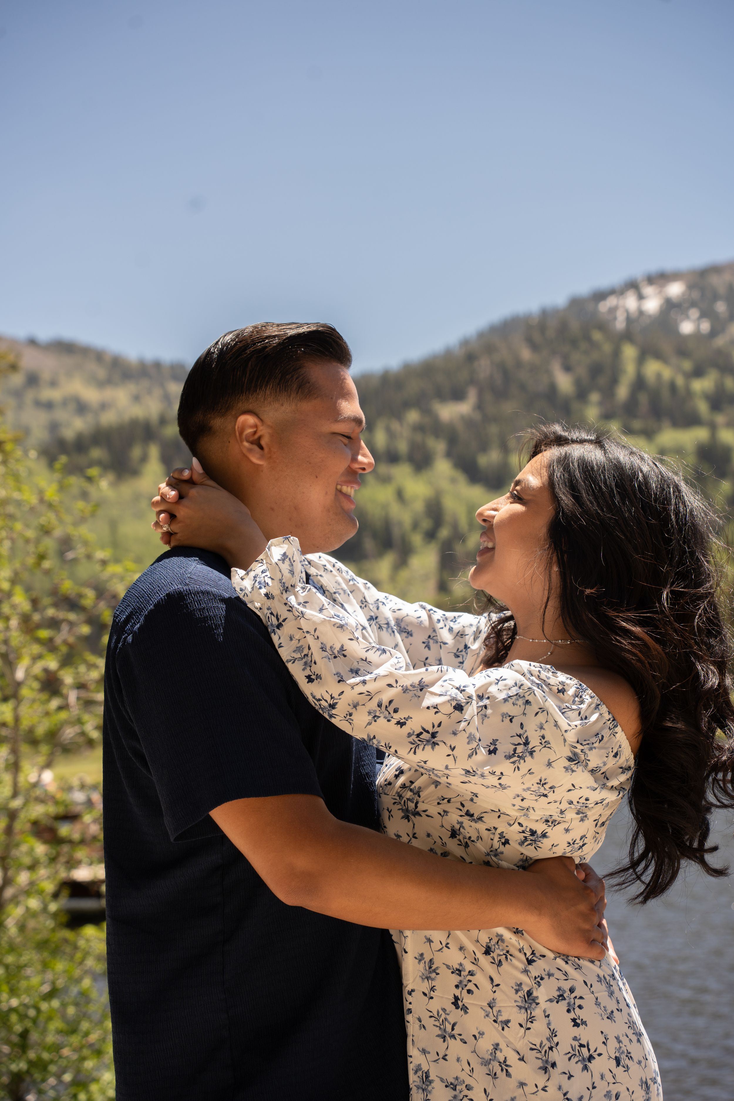 Couple embracing and smiling at each other with silver lake and mountains in background on sunny day.