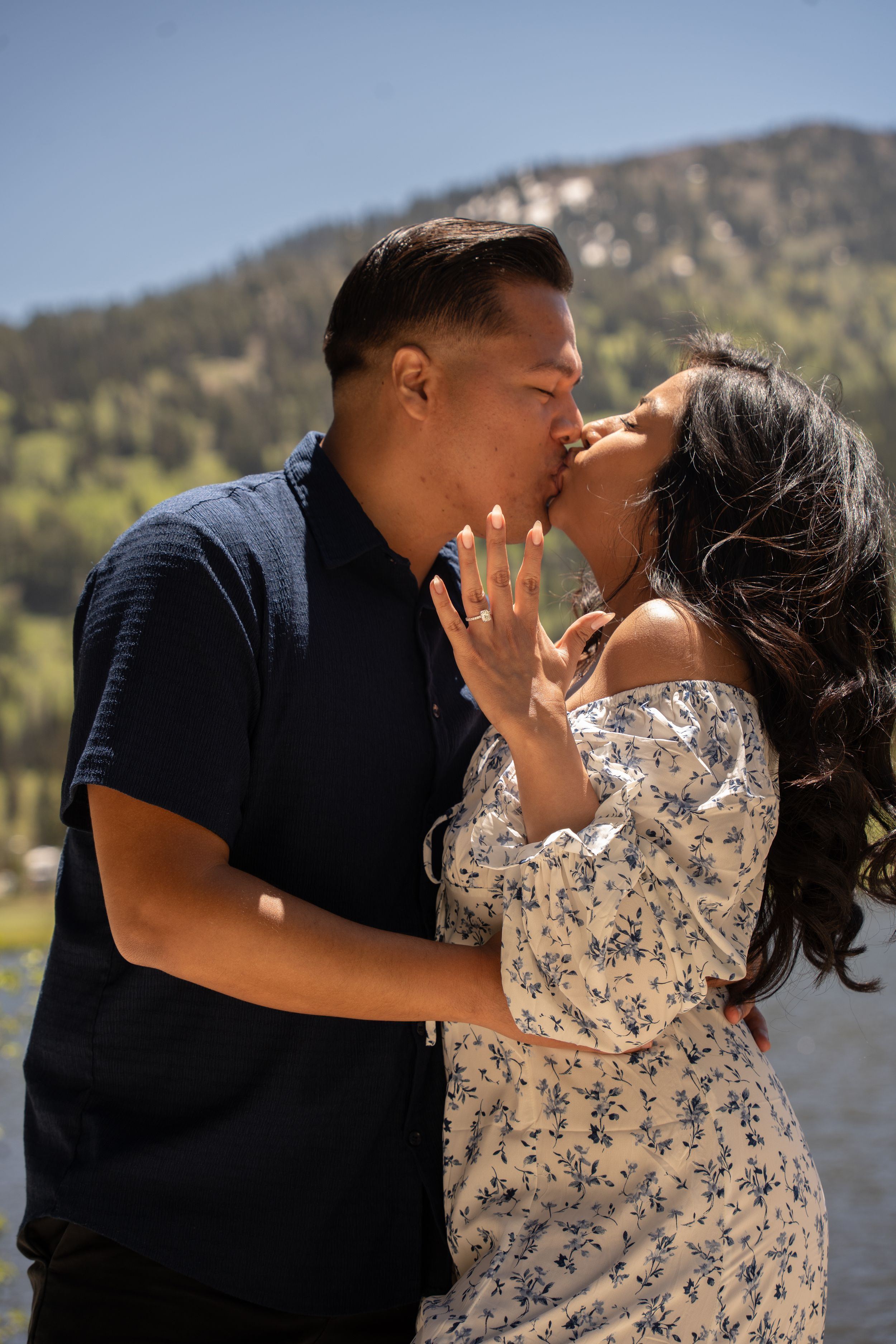 Couple sharing a romantic kiss outdoors with silver lake backdrop, woman showing engagement ring.