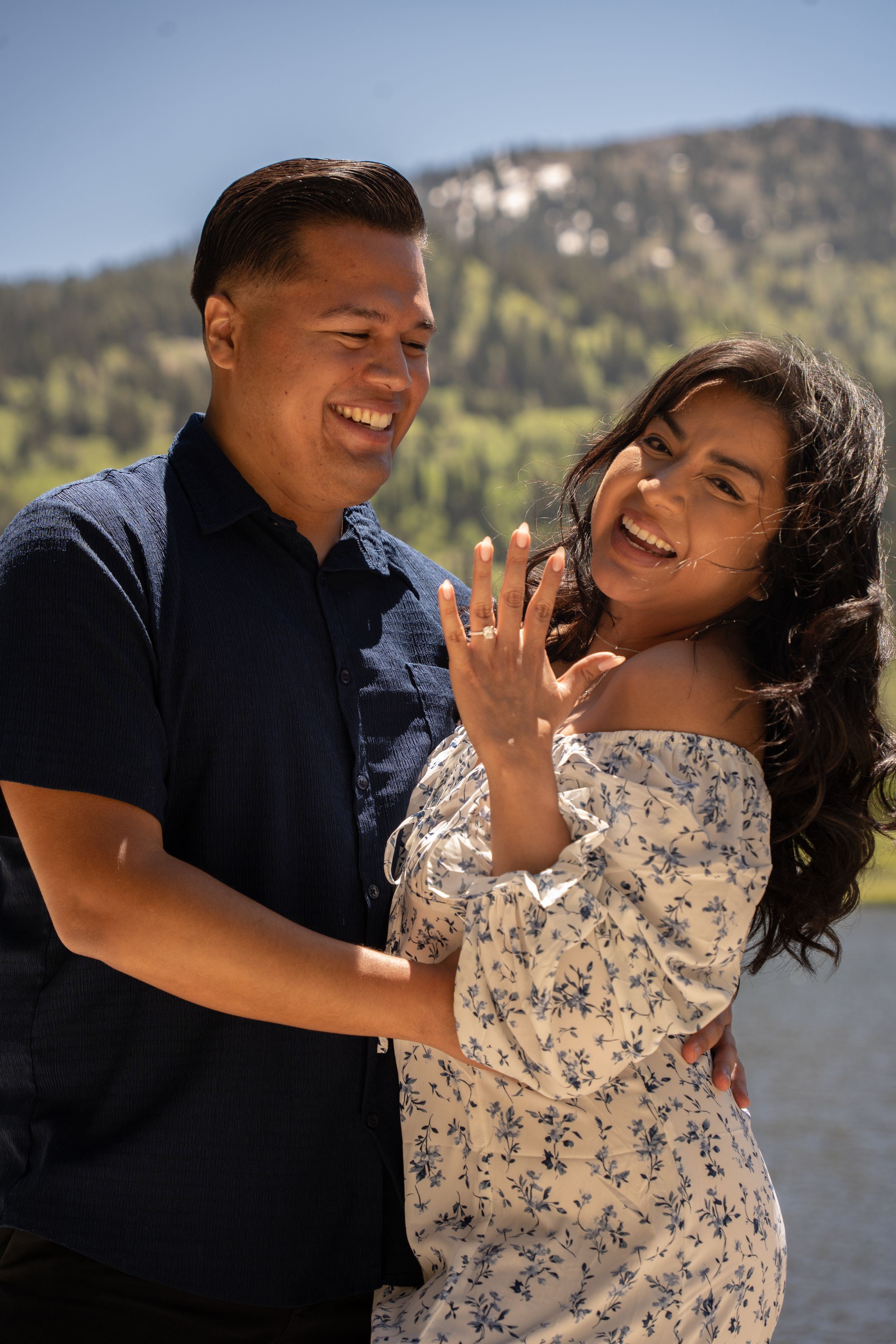 Happy couple celebrating engagement outdoors, woman showing ring, mountains and silver lake in background