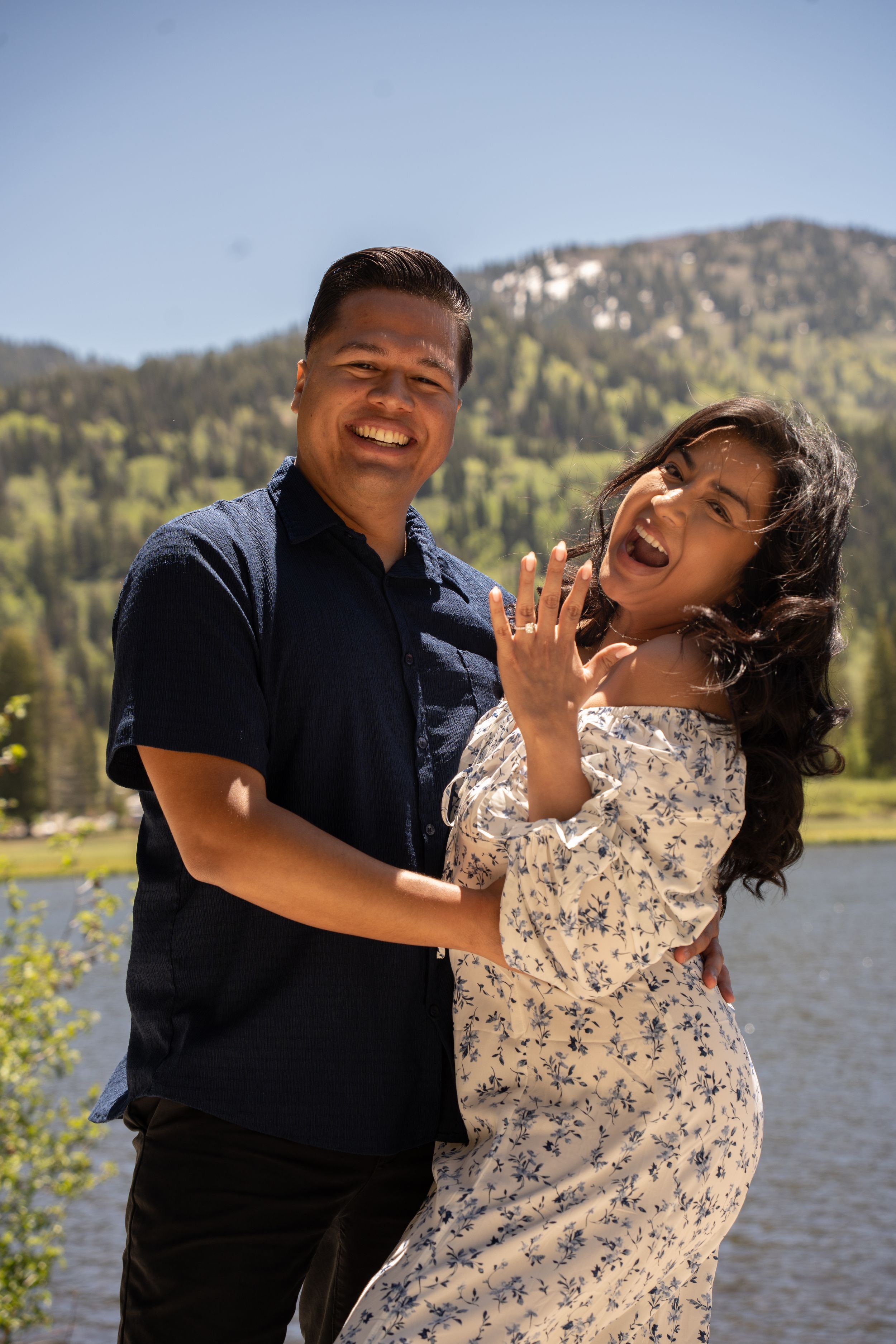 Happy couple celebrating engagement by a mountain lake, woman showing off ring, smiling joyfully outdoors.
