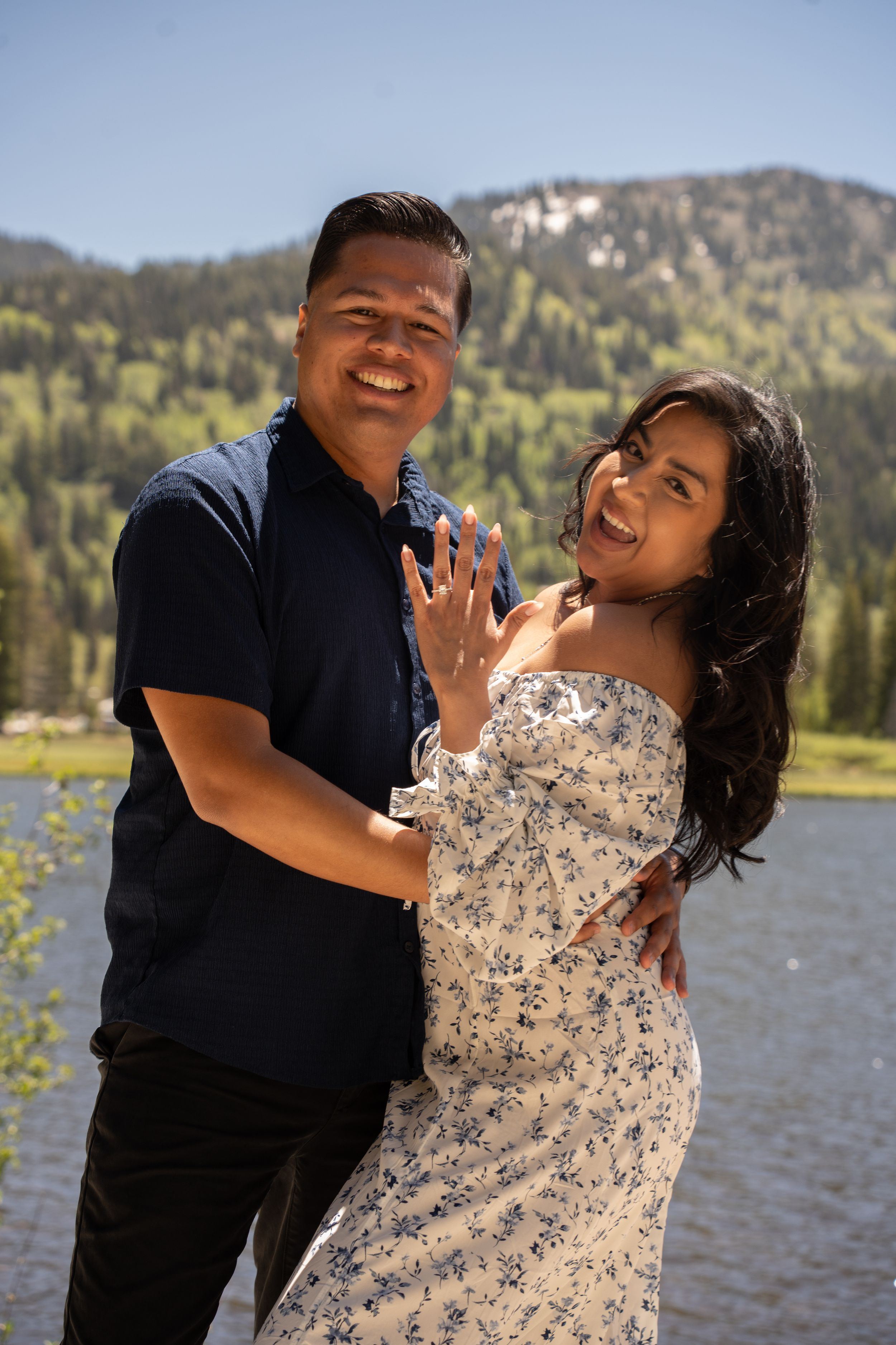Happy couple posing outdoors near a mountain lake, woman showing off engagement ring, surrounded by lush green trees.