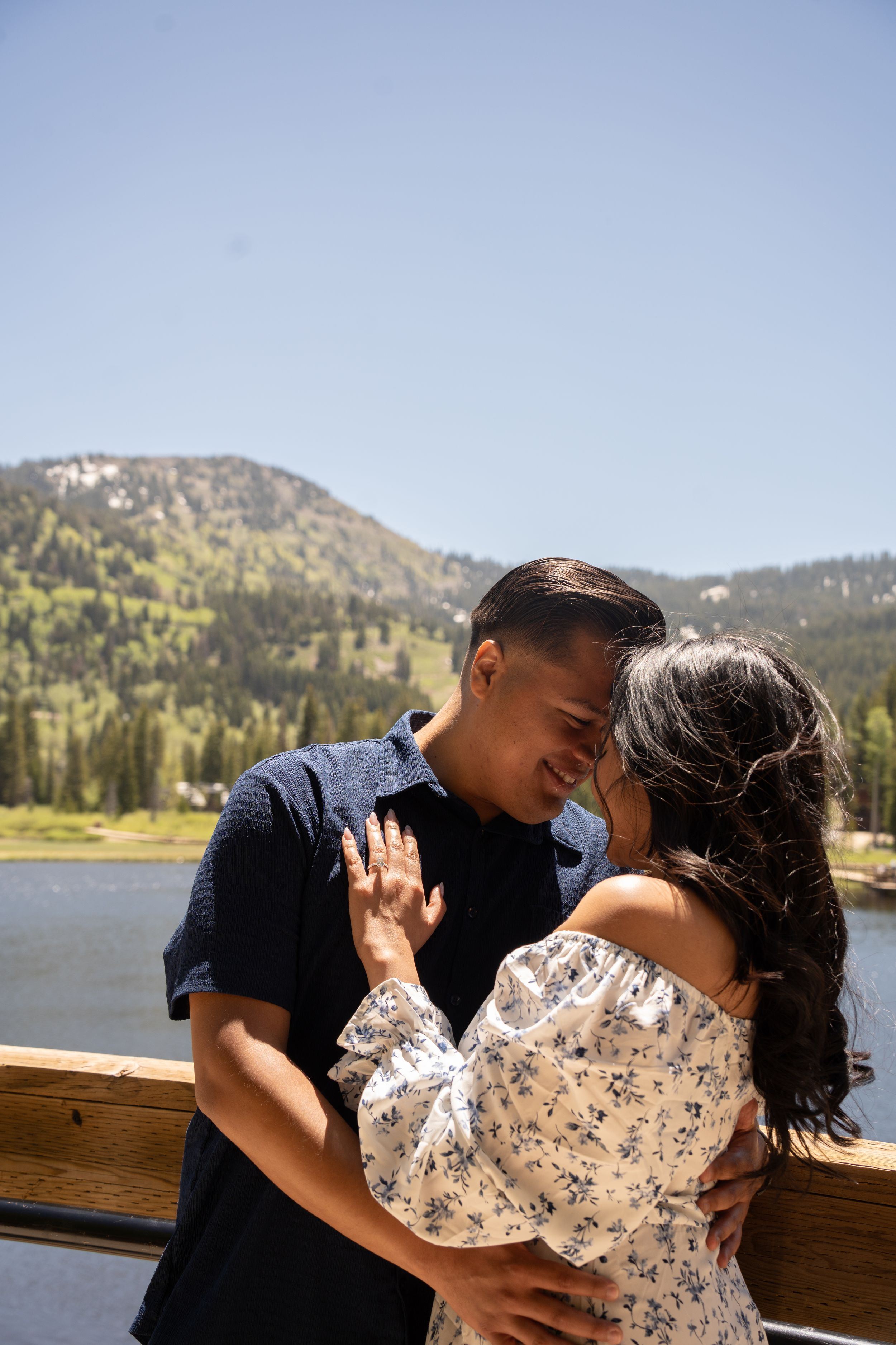 A couple shares a romantic moment on a wooden deck with a scenic mountain lake and pine trees in the background.