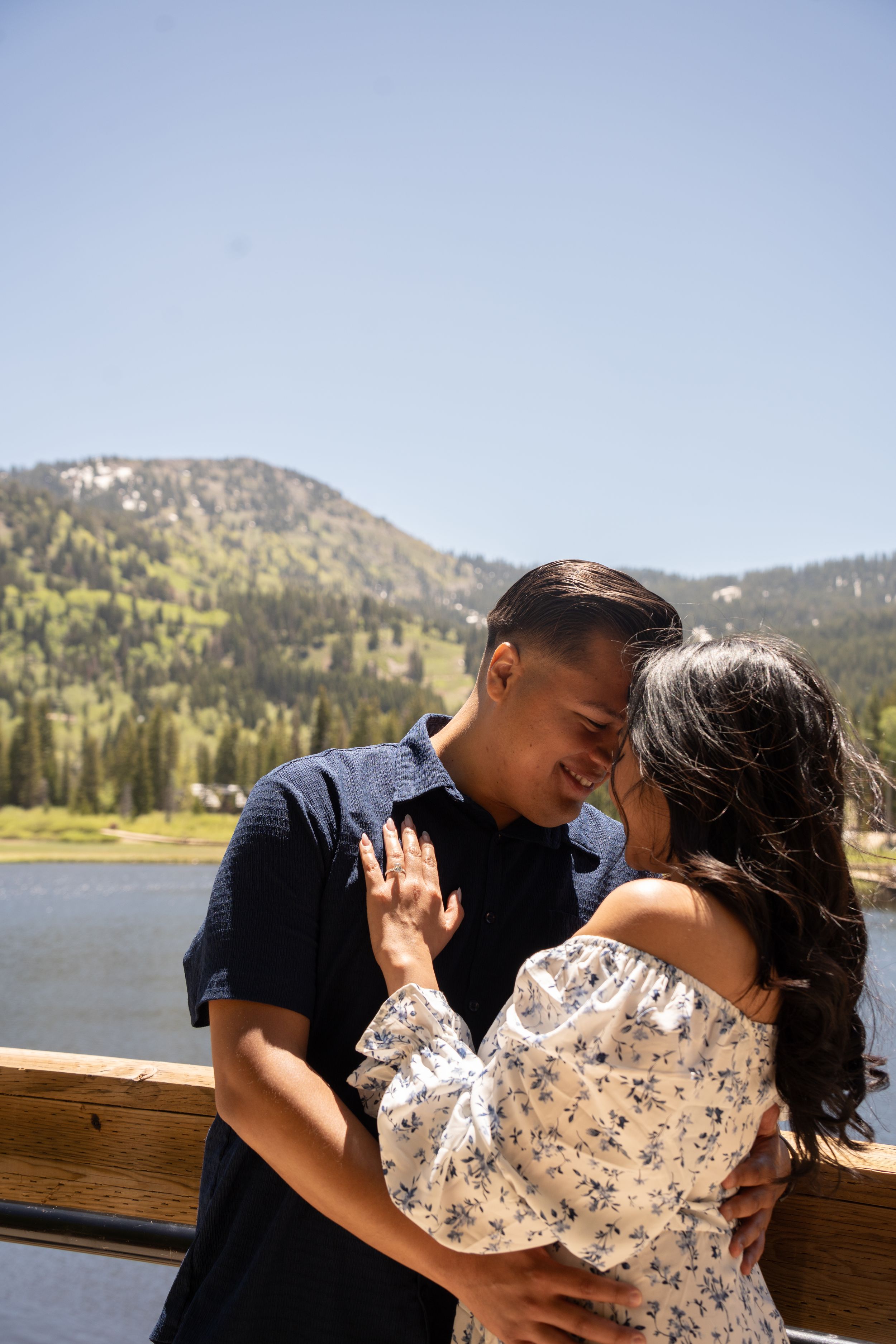 Couple embracing on a wooden bridge with a silver lake brighton and pine forest backdrop on a sunny day.