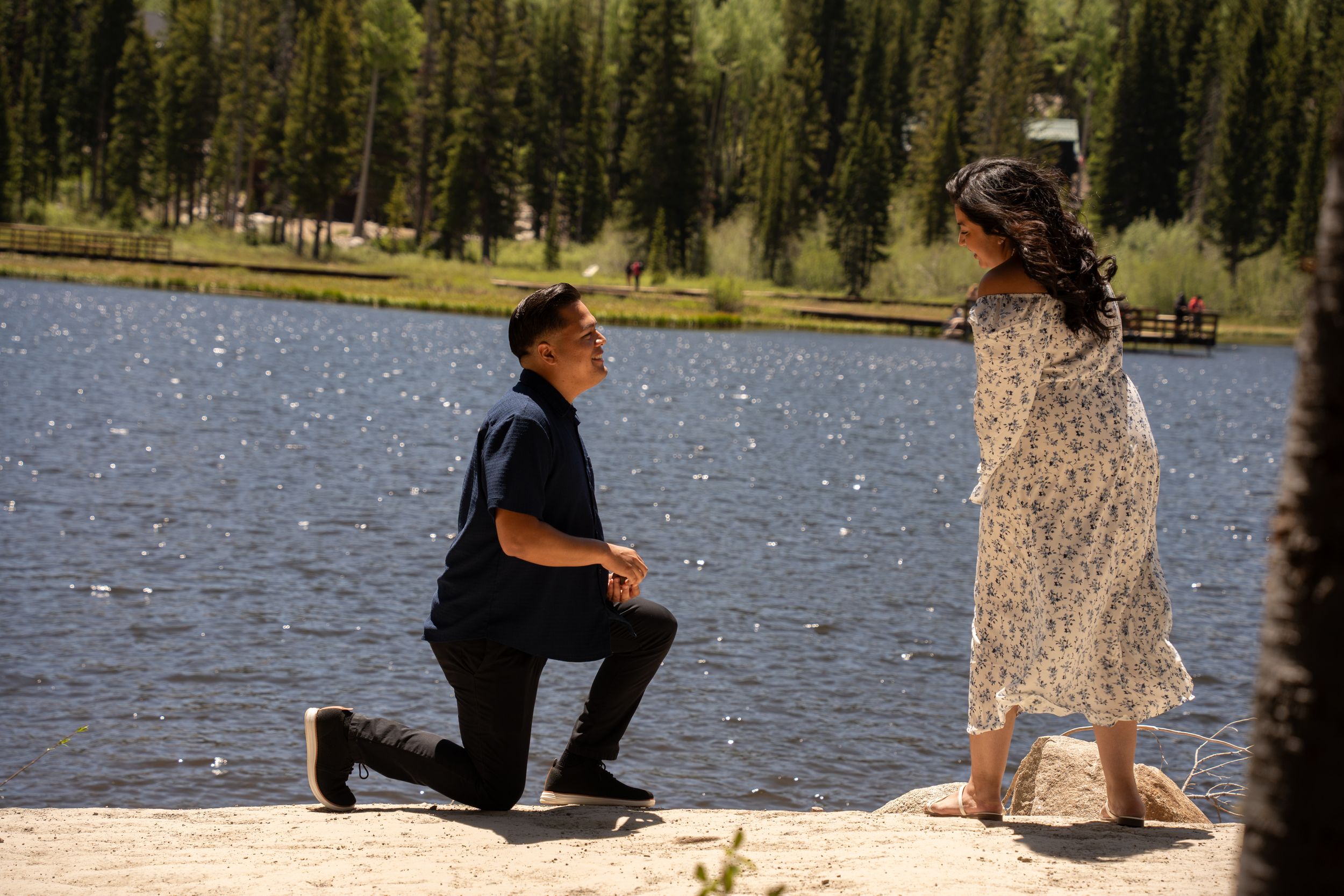 Man proposing on one knee to woman in floral dress by a serene silver lake surrounded by pine trees.