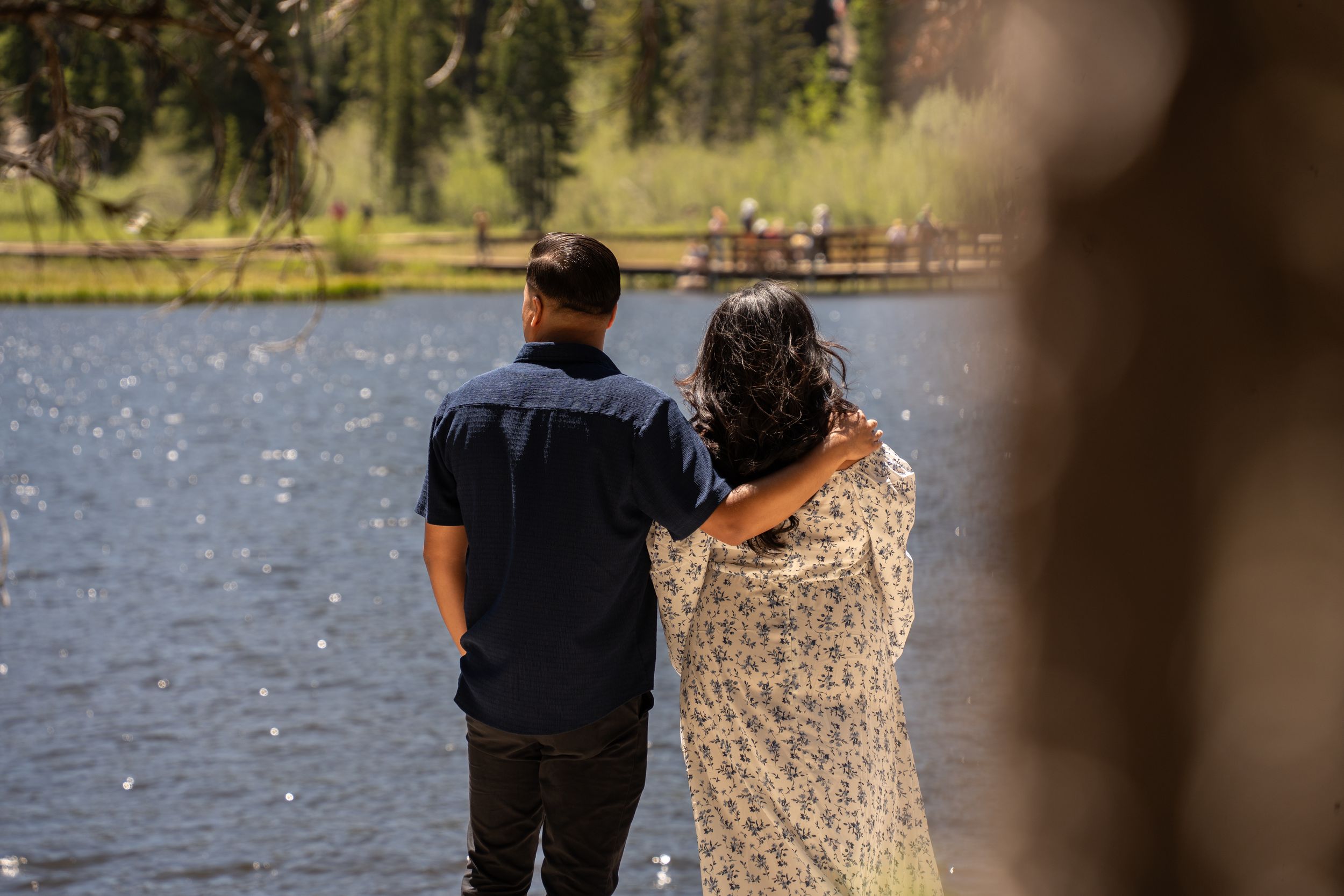 Couple standing arm in arm by a serene lake surrounded by lush green trees on a sunny day.