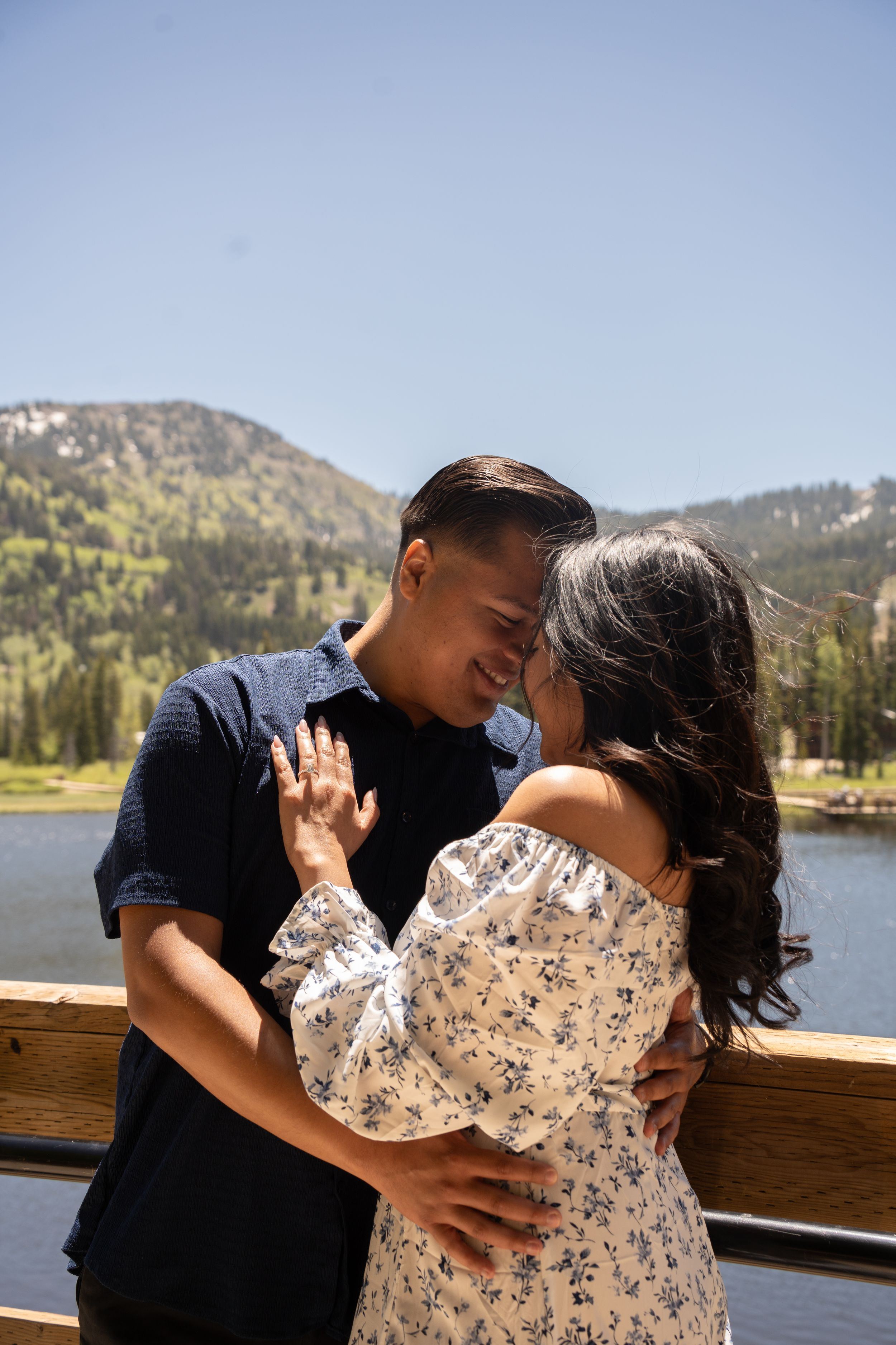 A couple embraces and touches foreheads romantically on a wooden deck with a mountain lake and forest backdrop of silver lake Utah.