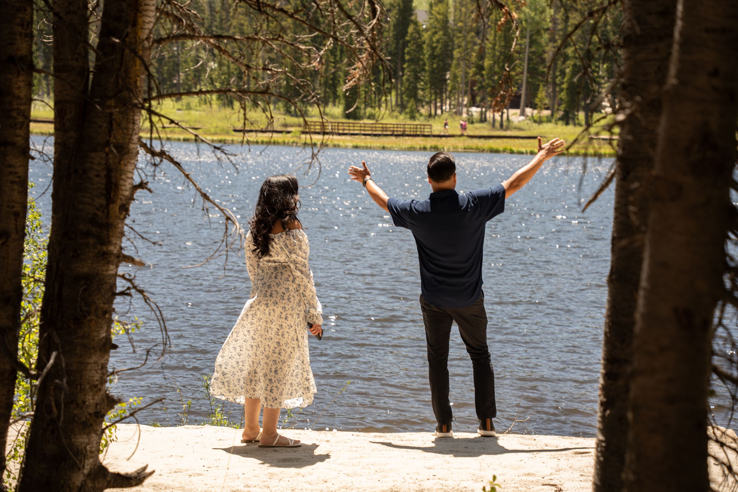 Couple standing by silver lake surrounded by pine trees, man raising arms in celebration.
