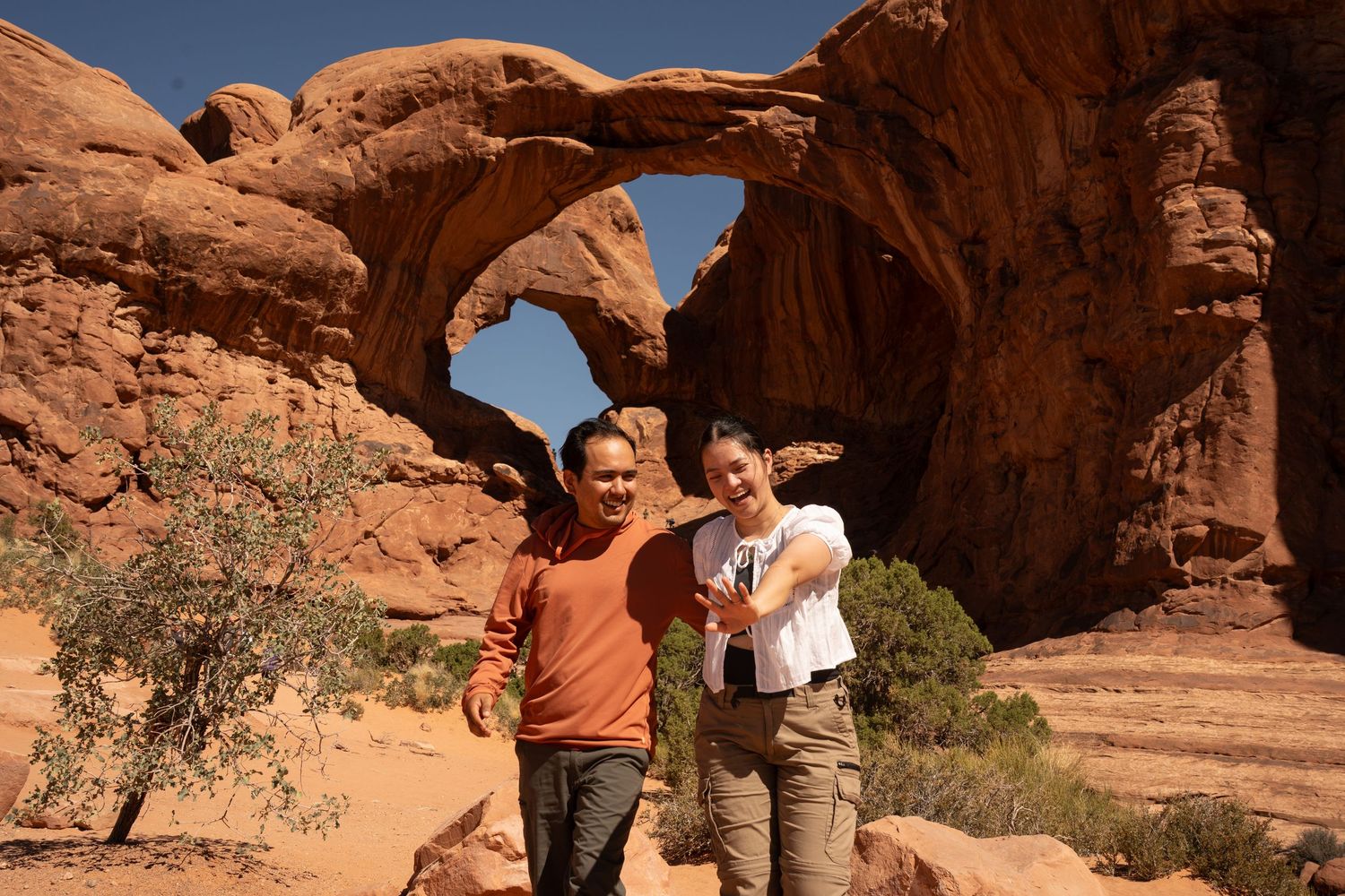 Two hikers pose and smile in front of a stunning natural sandstone arch at Arches National Park, Utah.