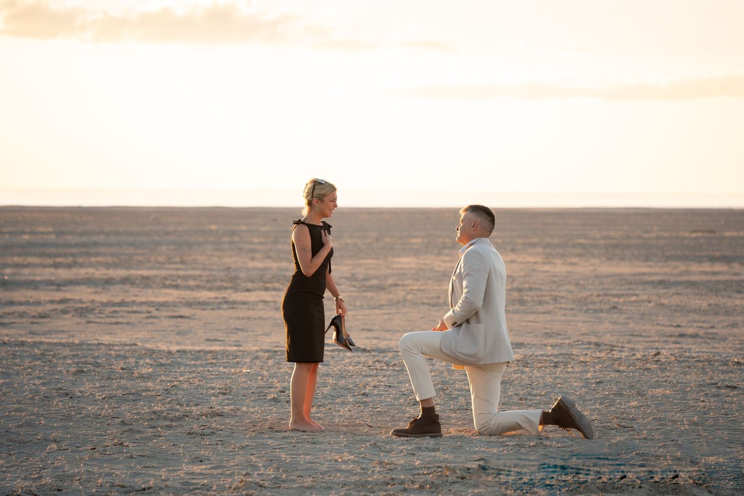 Man proposing on one knee to woman in black dress at sunset on a vast desert landscape at salt flats
