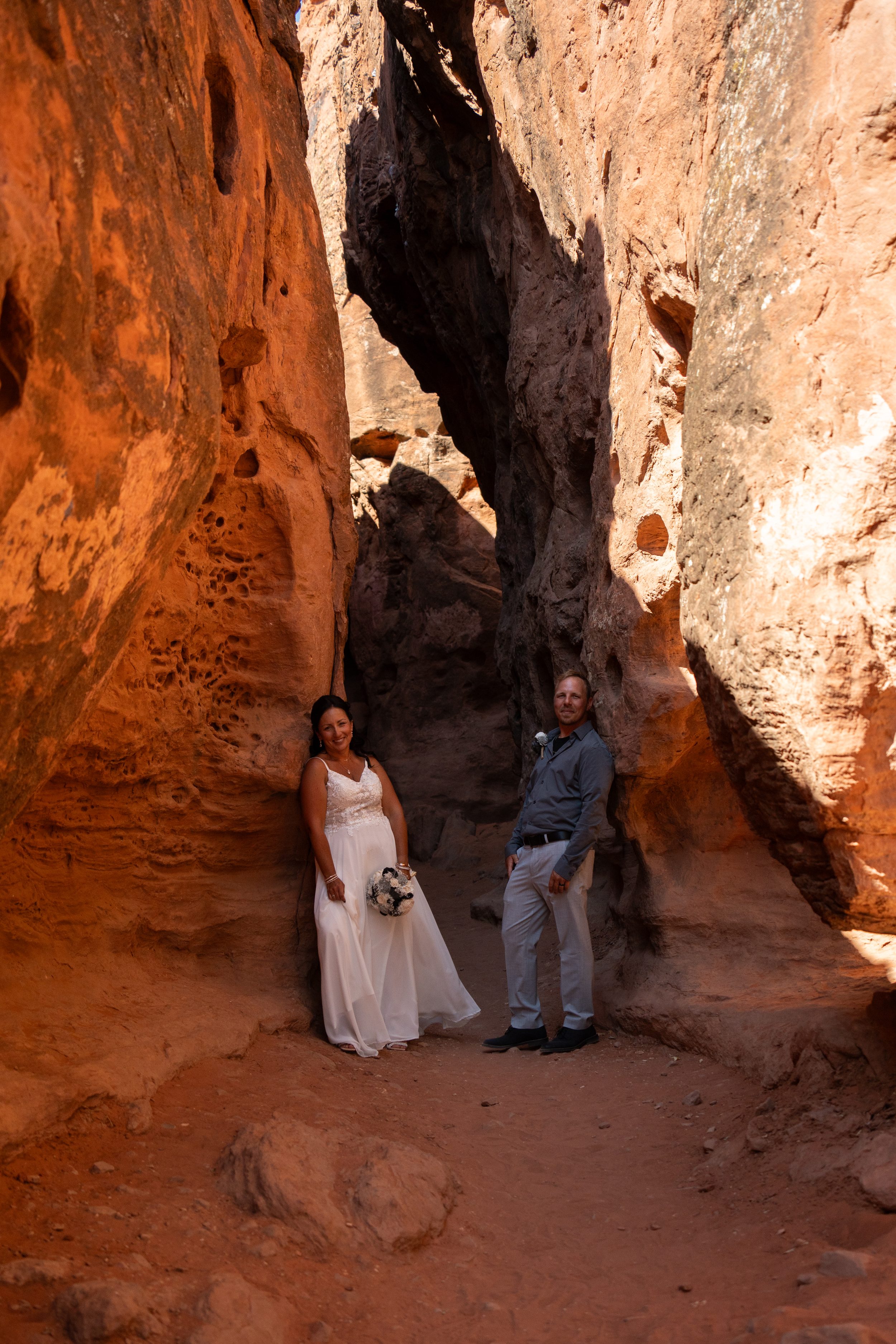 Bride and groom standing together in a shaded red rock alcove at Pioneer Park, holding a black and white bouquet.