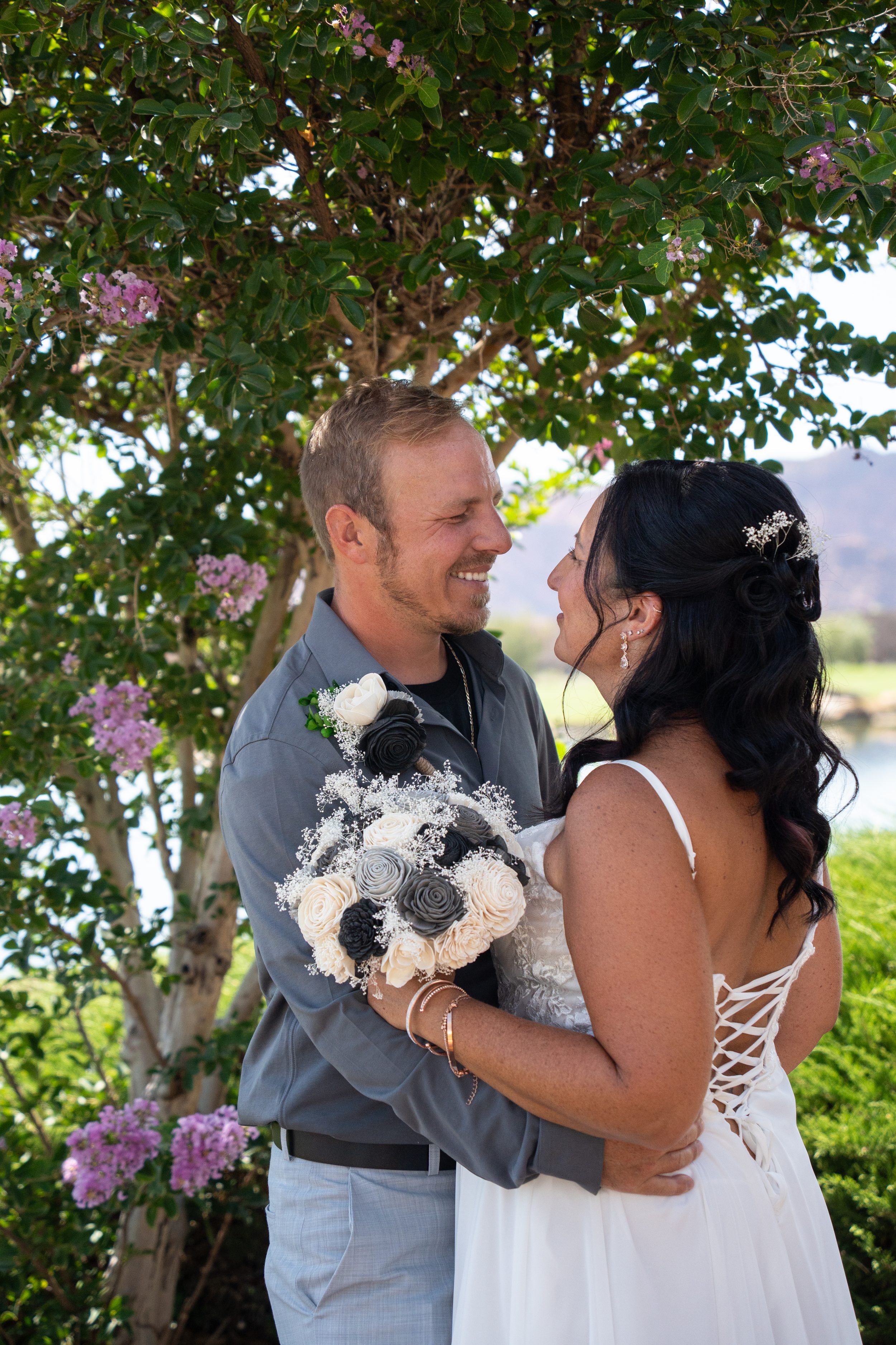 Bride and groom embracing under a flowering tree with a lake and mountains behind them at The Ledges.
