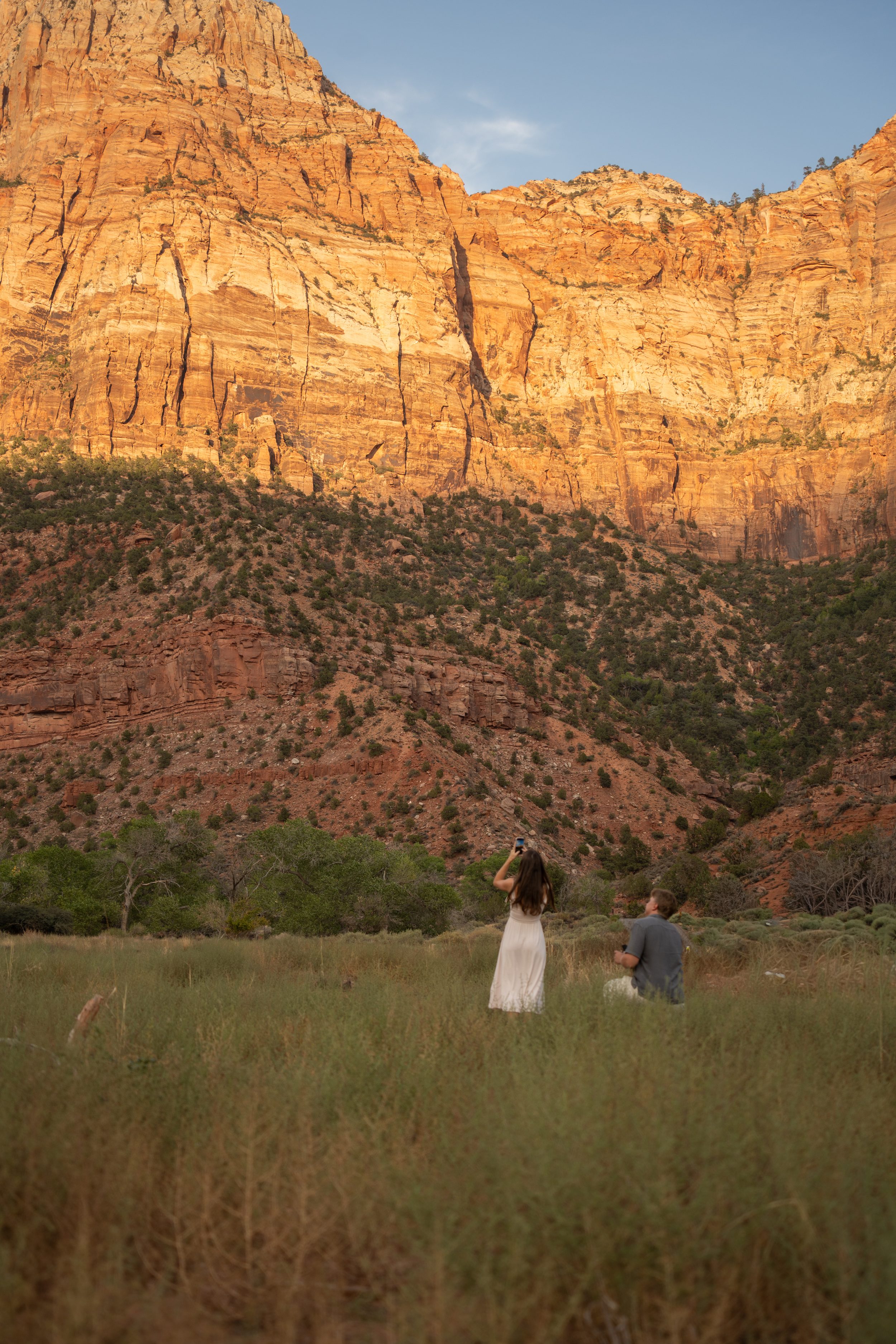 Lyndsey photographing the meadow on her phone, unaware Jack is kneeling behind her with the ring.