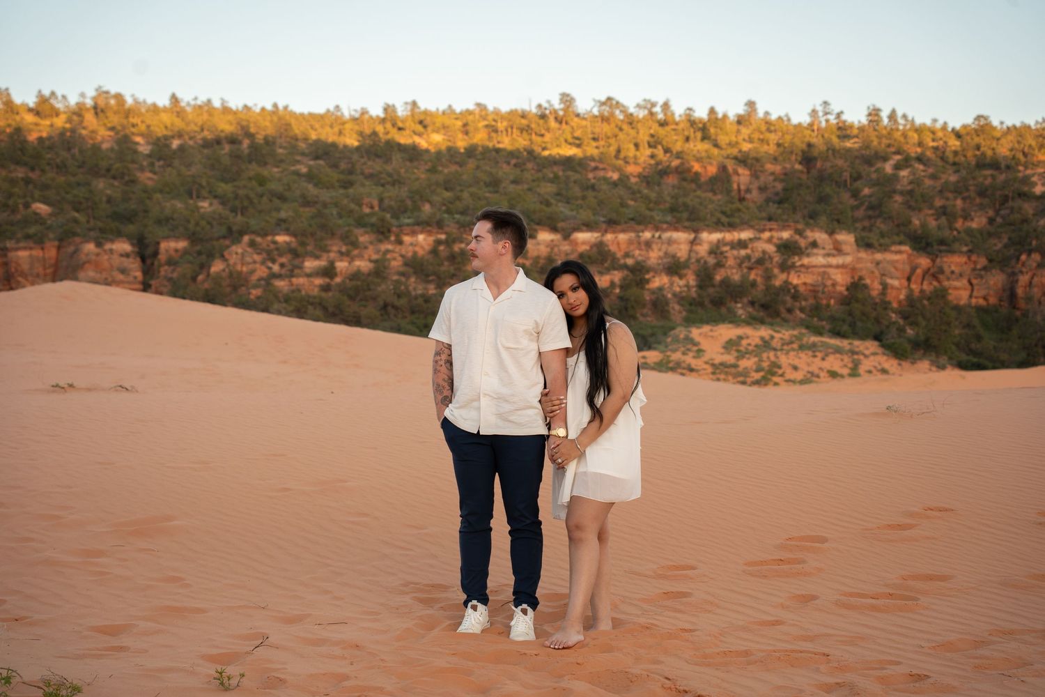 engaged Couple holding hands on red sand dunes with sandstone cliffs and green trees at golden hour.