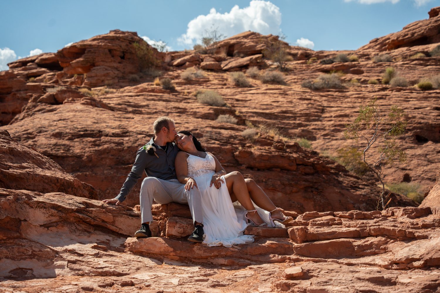 Bride in white and groom in neutral gray sitting together on red rock, an example of coordinating rather than matching outfits