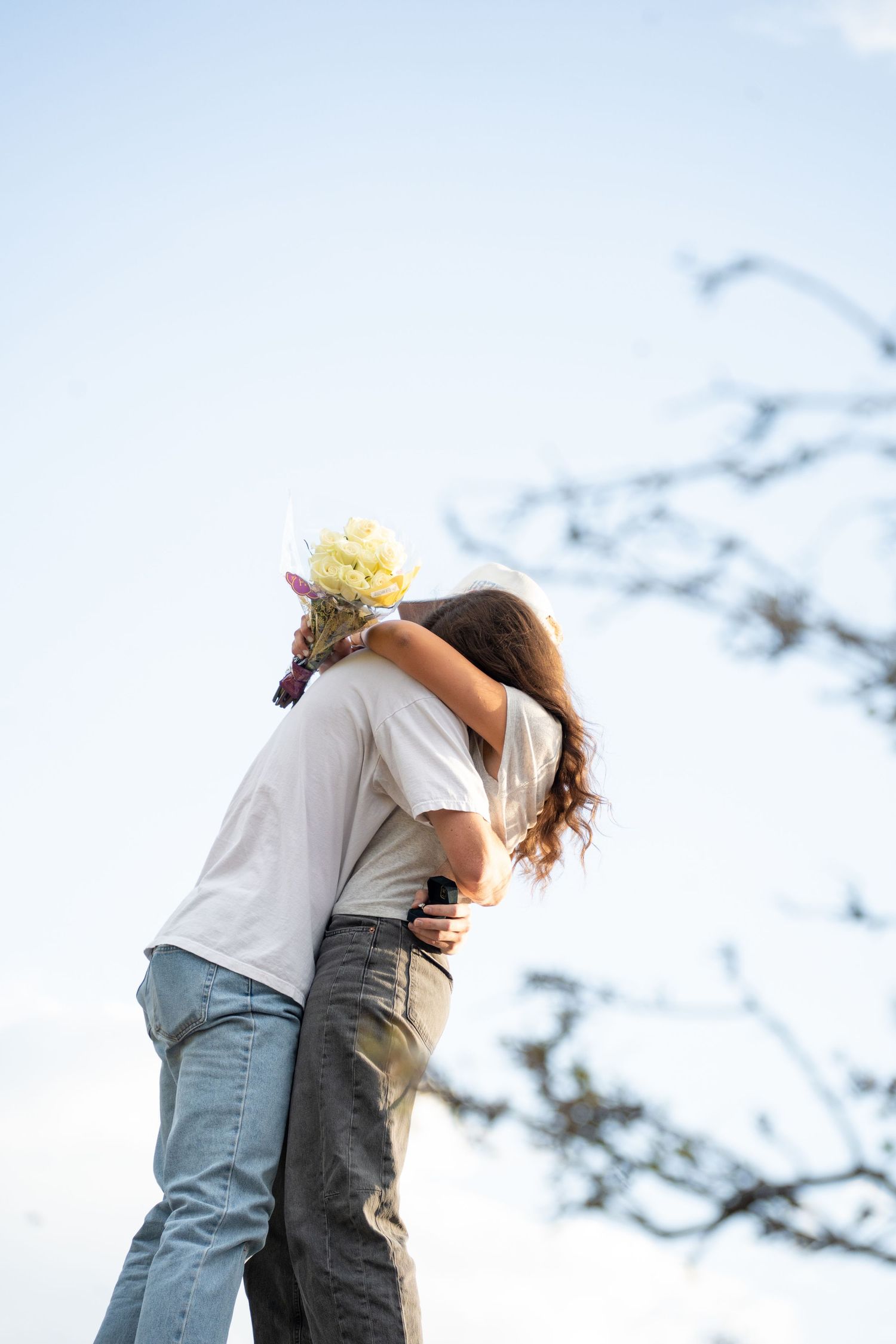 A newly engaged couple embraces outdoors against a bright blue sky with tree branches visible in the background. Lindon Utah