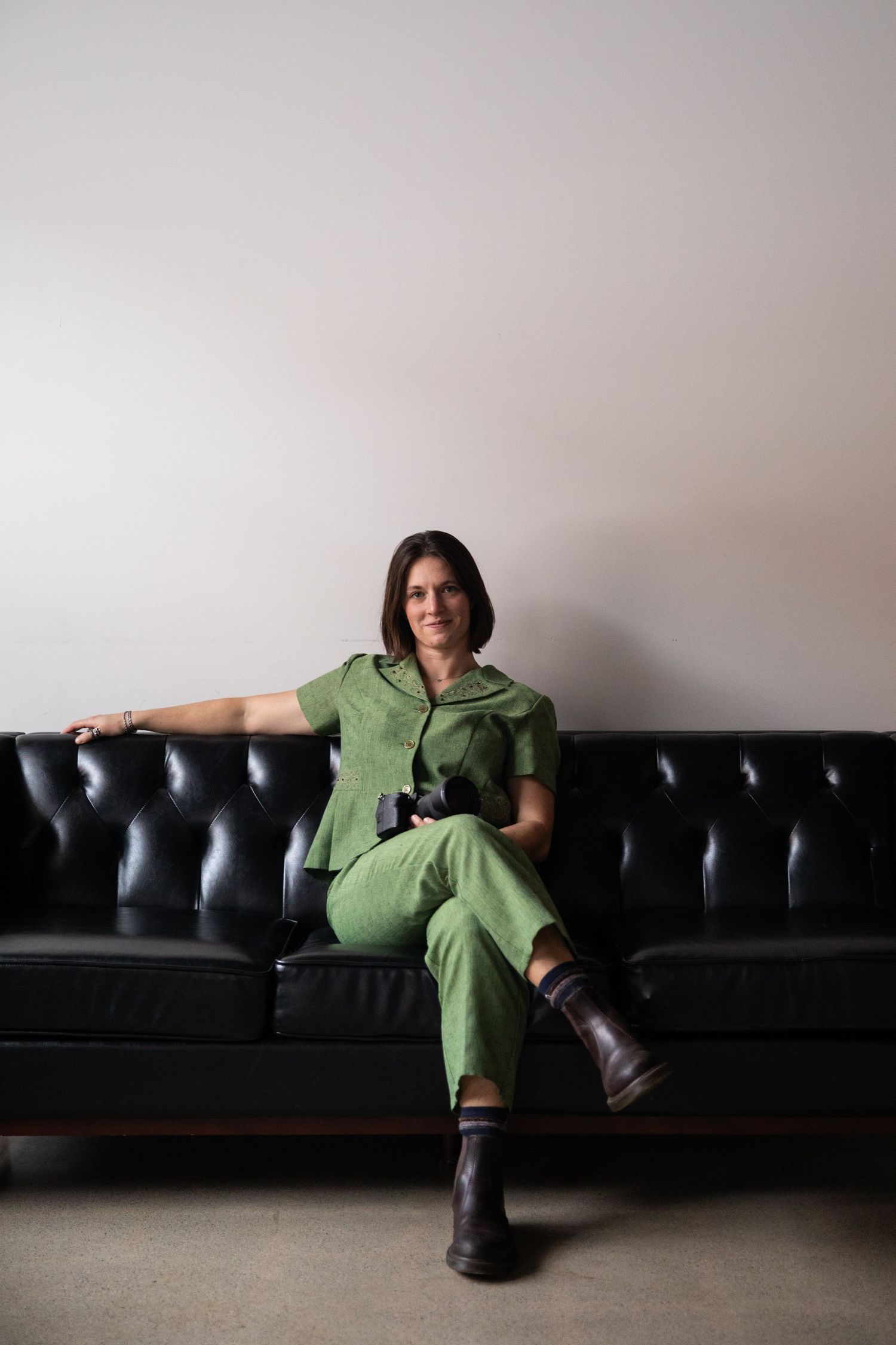 A wedding photographer in a stylish green jumpsuit relaxes on a black leather couch against a light gray wall.