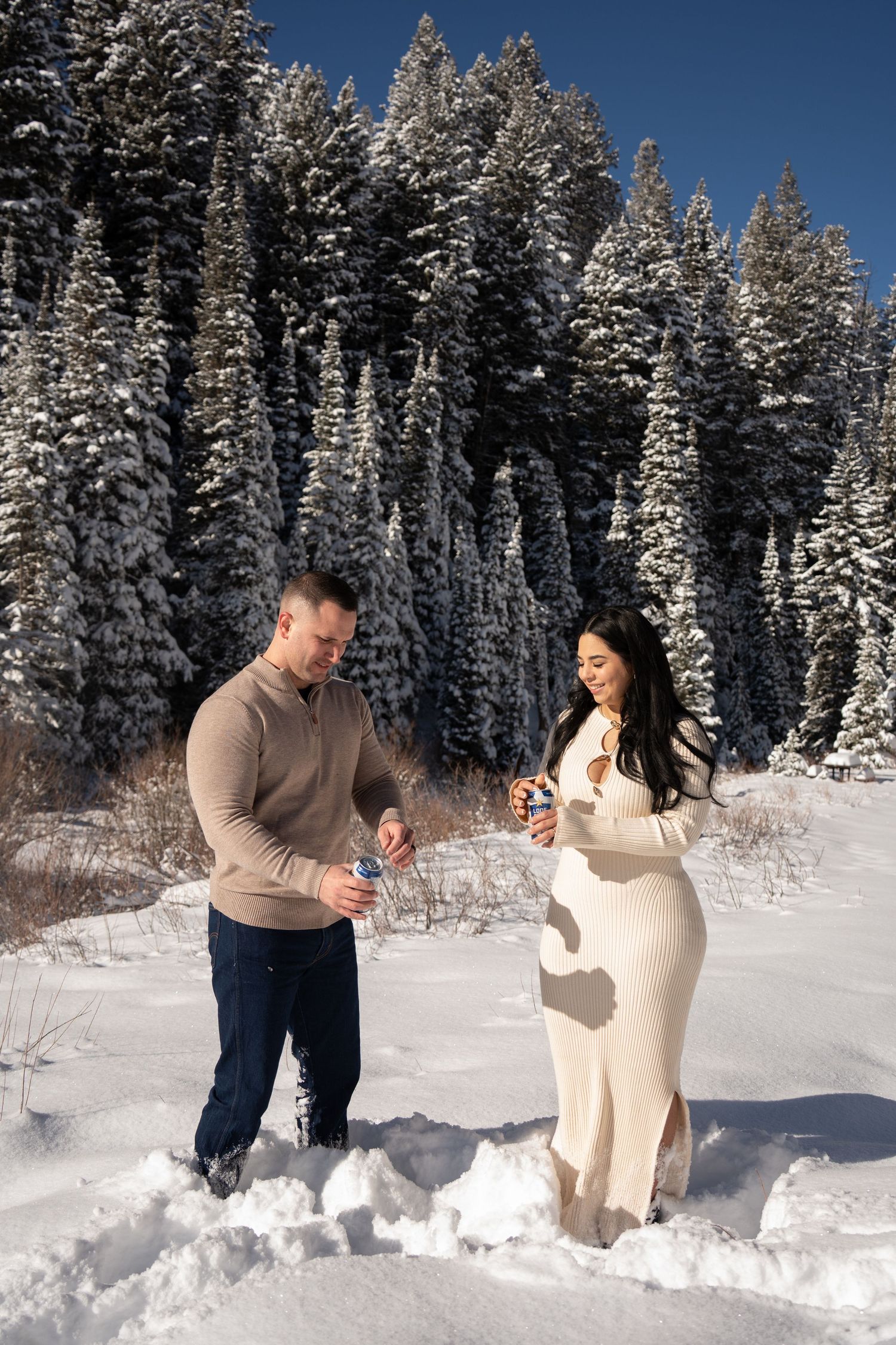 Couple in snowy winter forest, woman in white dress and man in beige sweater, opening champagne in fresh snow.