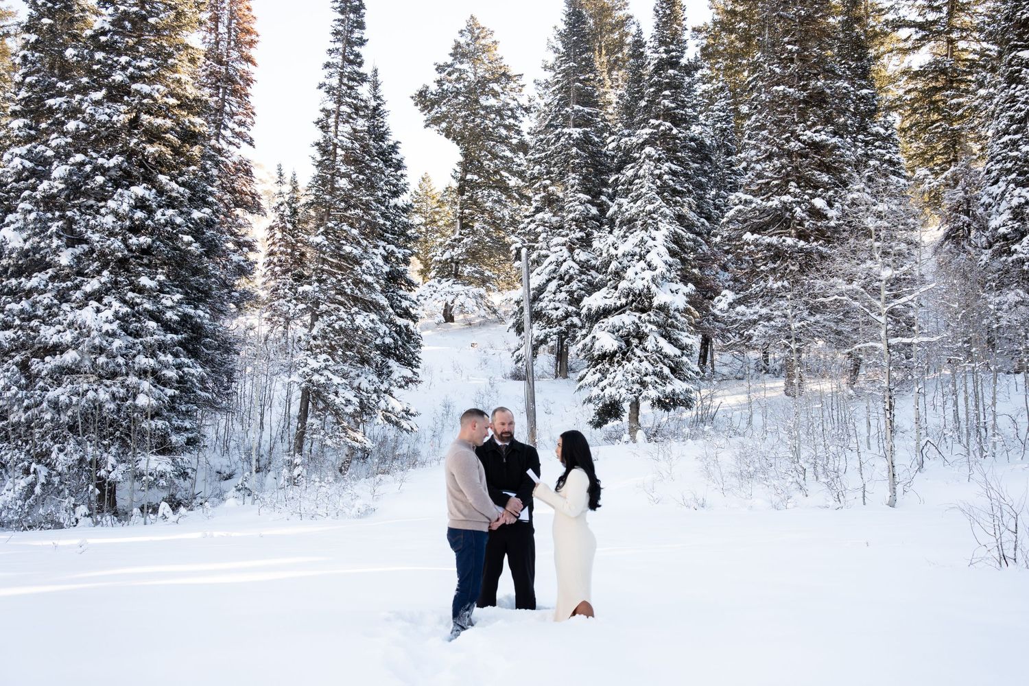 Couple holding hands in snowy winter forest with tall pine trees covered in snow during an outdoor elopement ceremony.
