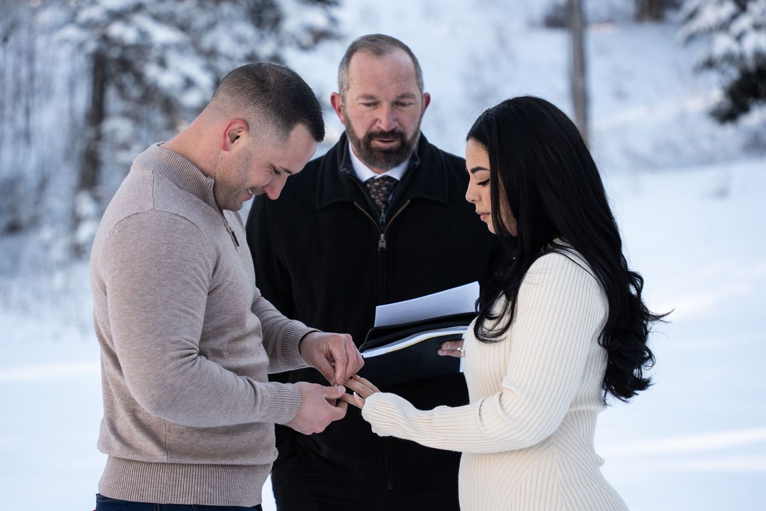 Couple exchanging rings during a winter wedding ceremony outdoors in snowy landscape with officiant.