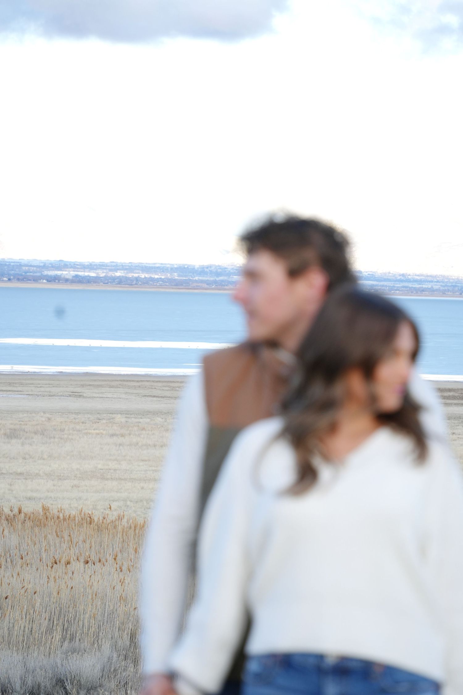 A couple stands together near a serene Great Salt Lake shore with dry grass fields under a cloudy sky
