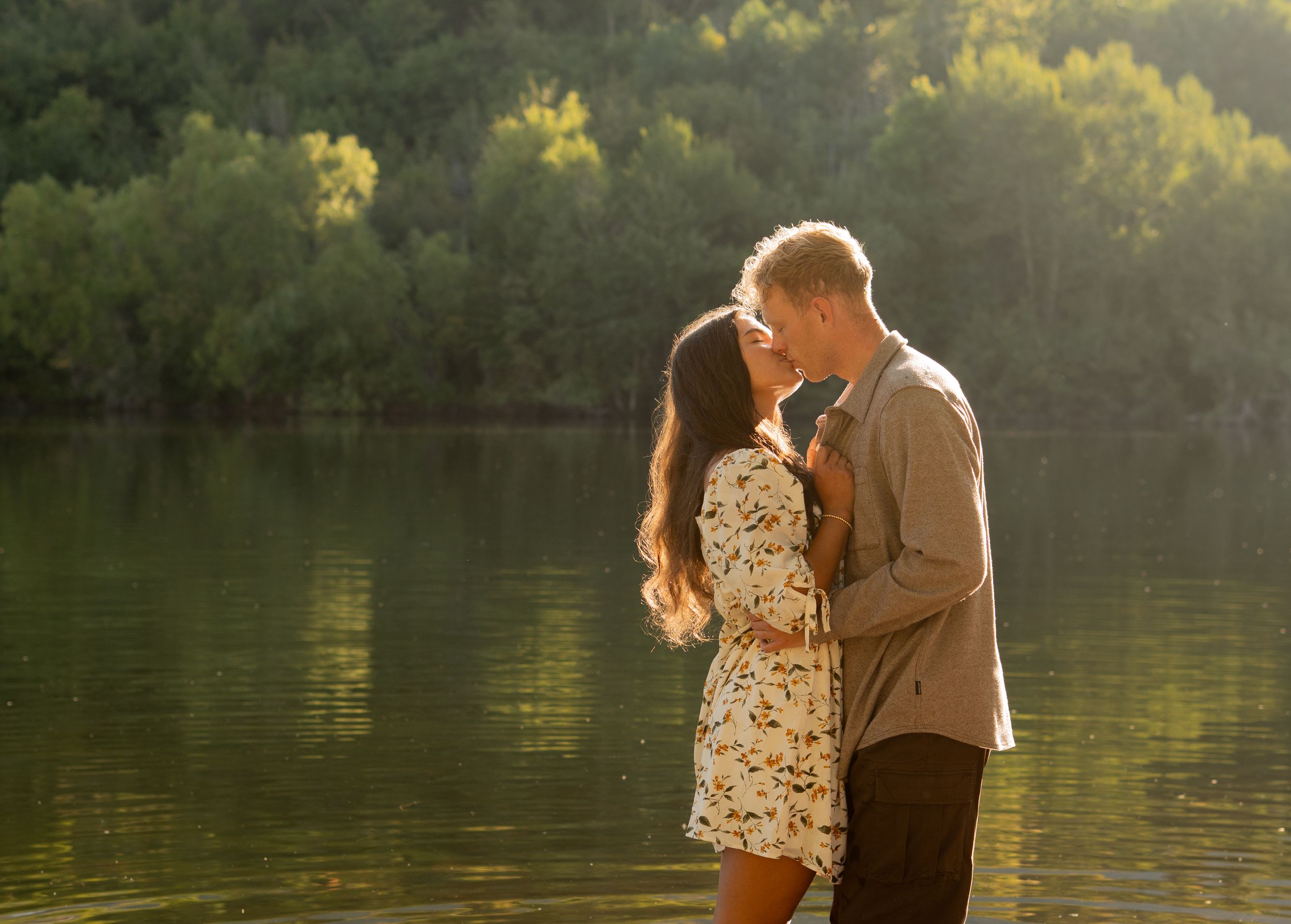Couple sharing a romantic kiss by a serene mountain lake in Sandy, Utah, surrounded by lush green trees in golden summer sunlight