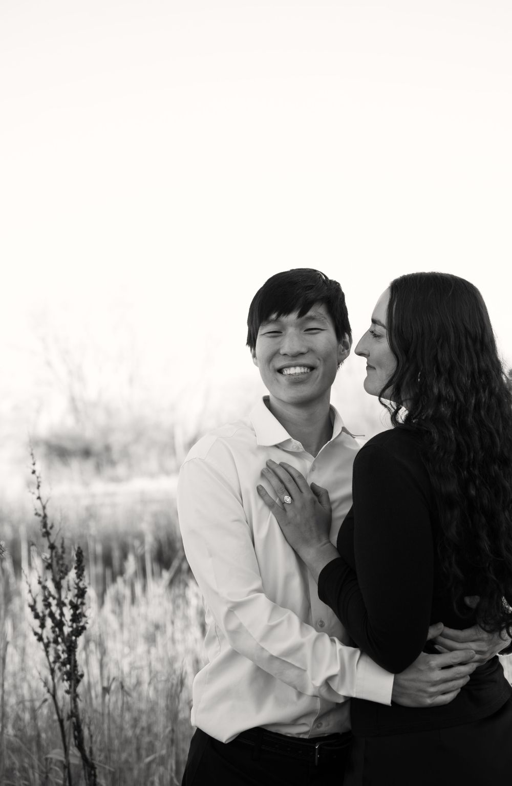 Black and white photo of a smiling man with a woman showing her engagement ring, standing together in tall grass