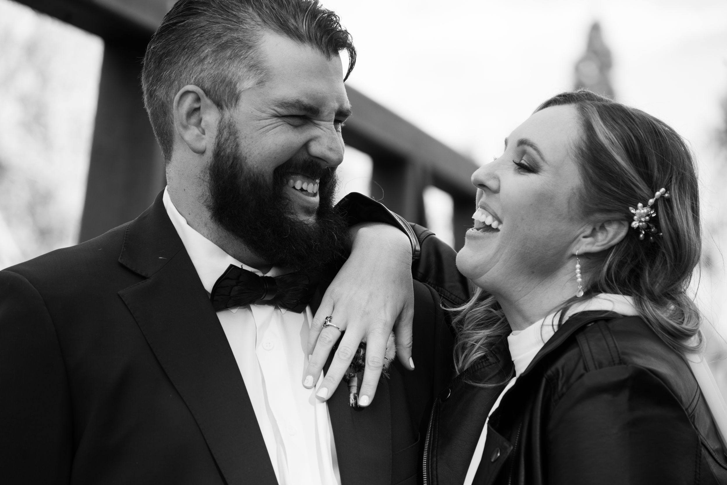 Black and white photo of a laughing couple in formal wedding attire, sharing a joyful moment together on a bridge
