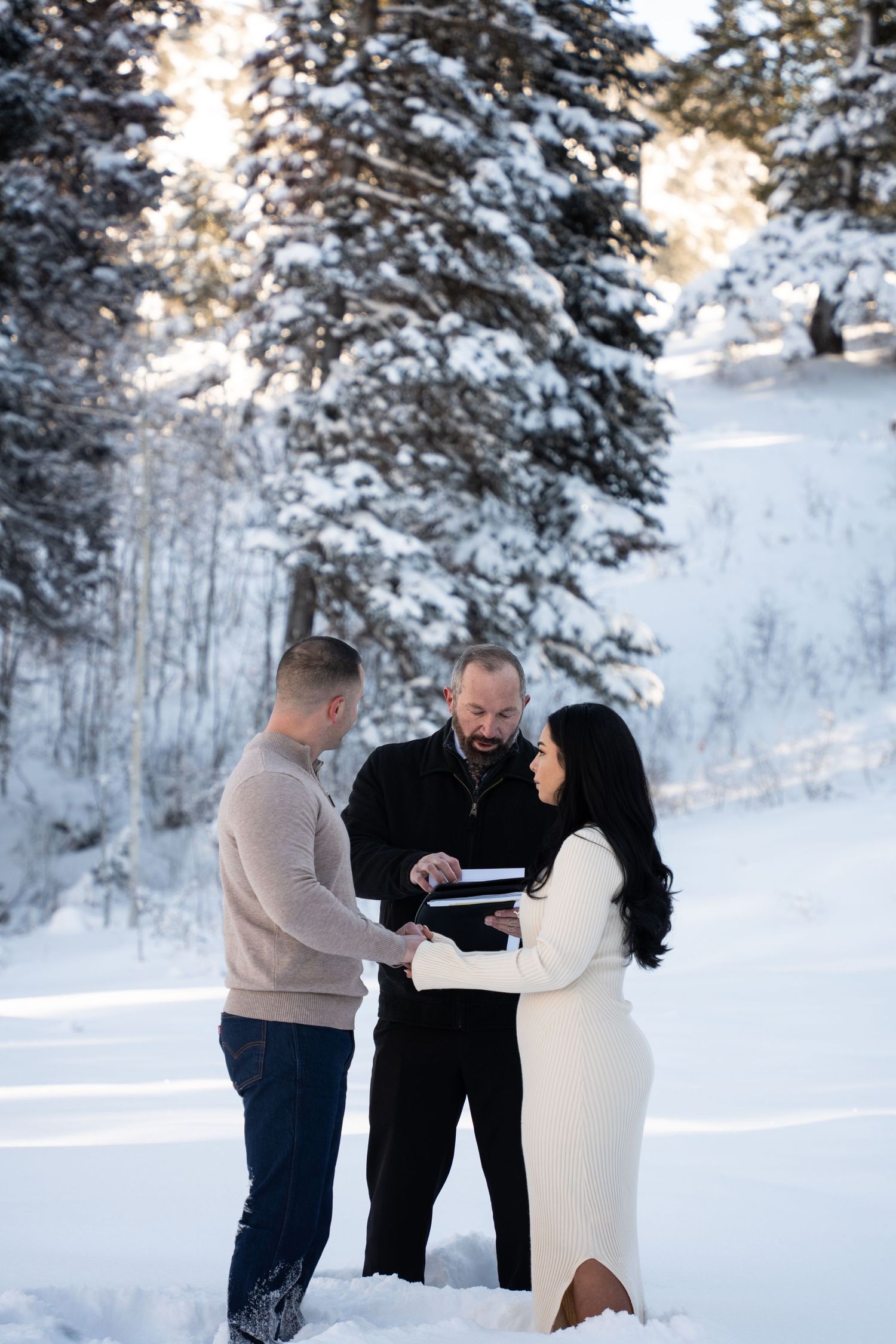 Couple exchanging vows in snowy winter outdoor ceremony with officiant, surrounded by snow-covered pine trees in Salt Lake, Utah