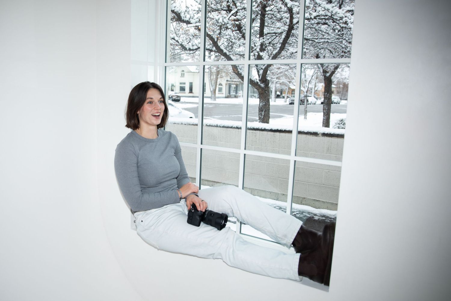 Photographer in gray sweater sits near snowy window while working on laptop in cozy winter setting.