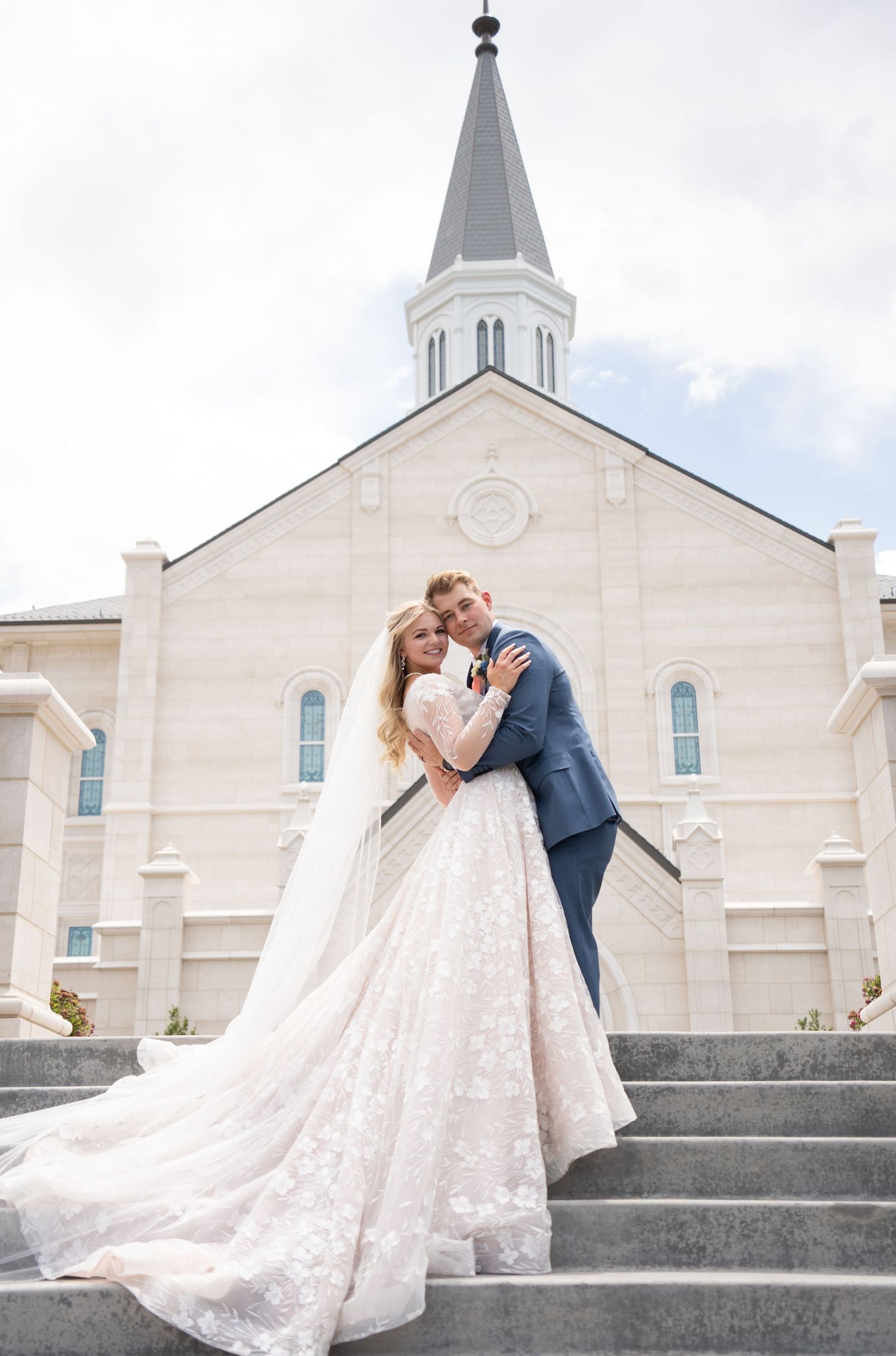 Bride and groom embrace on Taylorsville Temple steps, bride in lace ballgown with veil, groom in blue suit, summer wedding