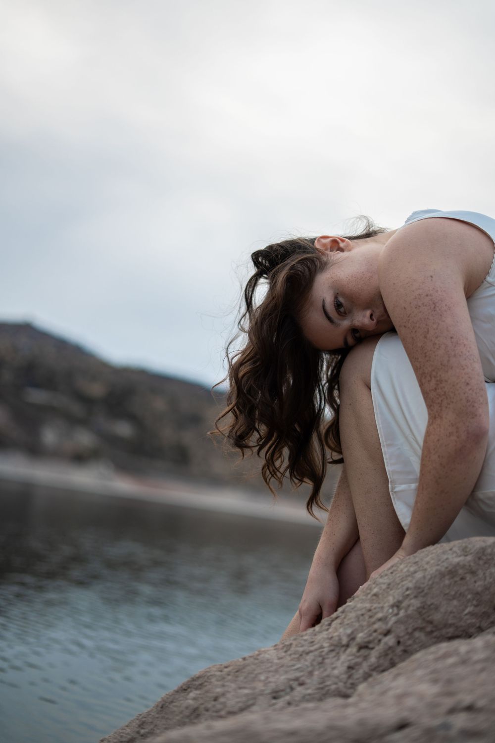 A young woman with freckles and dark wavy hair sits on rocks