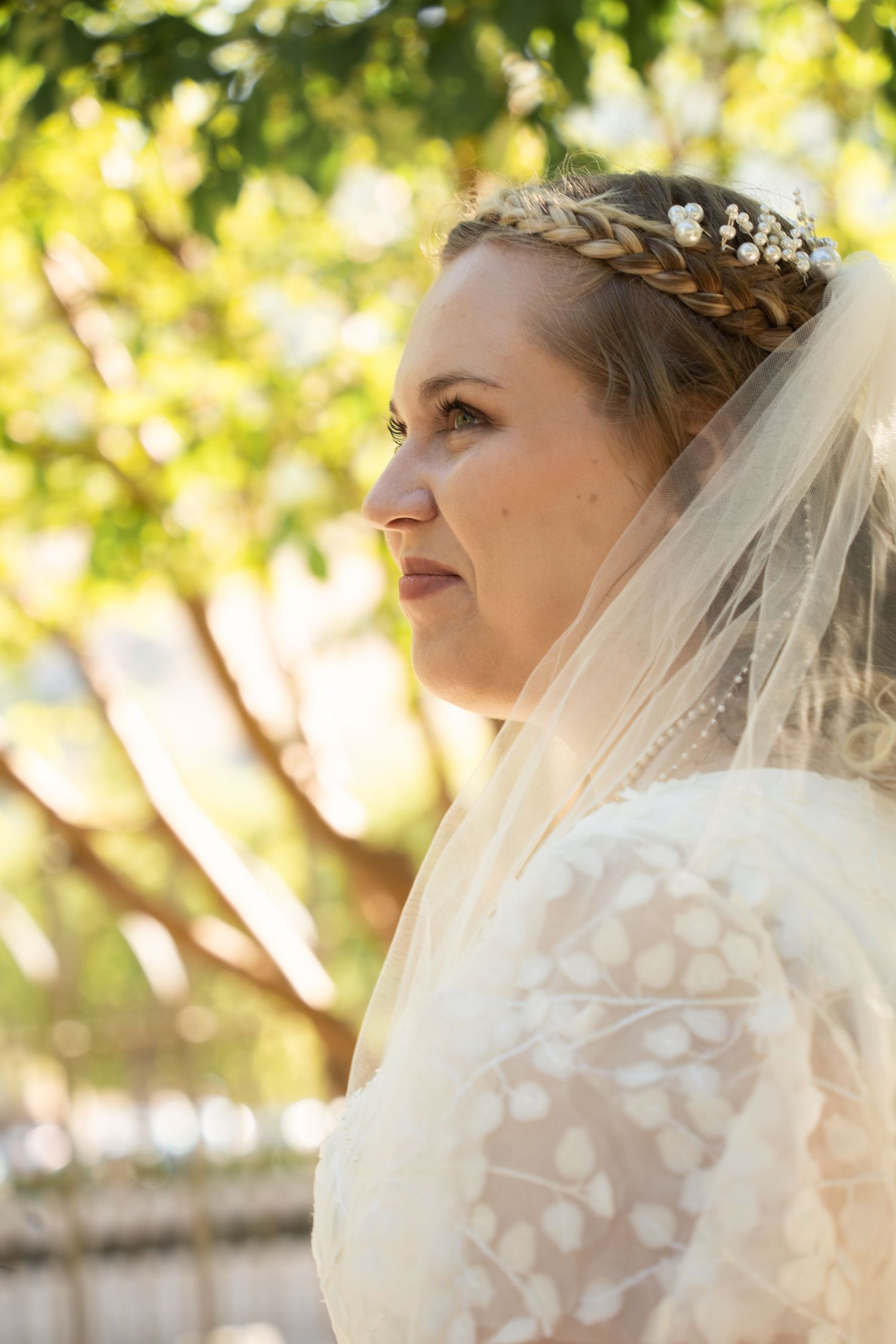 Bride in lace dress and veil with braided crown and floral headpiece, gazing serenely outdoors near green trees at Draper Temple