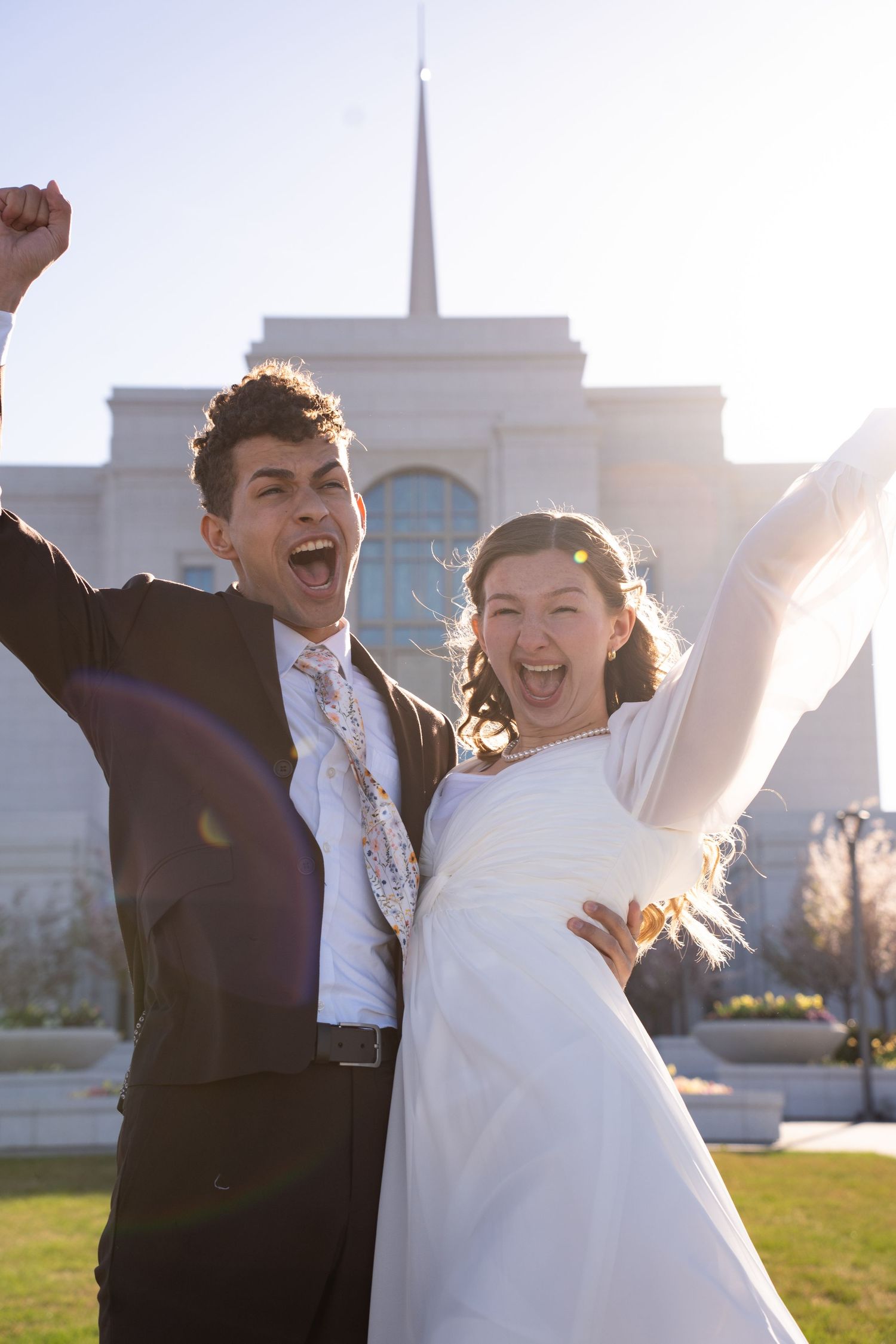 Joyful newlyweds celebrating outside Jordan River temple, bride in white gown and groom in dark suit with arms raised.