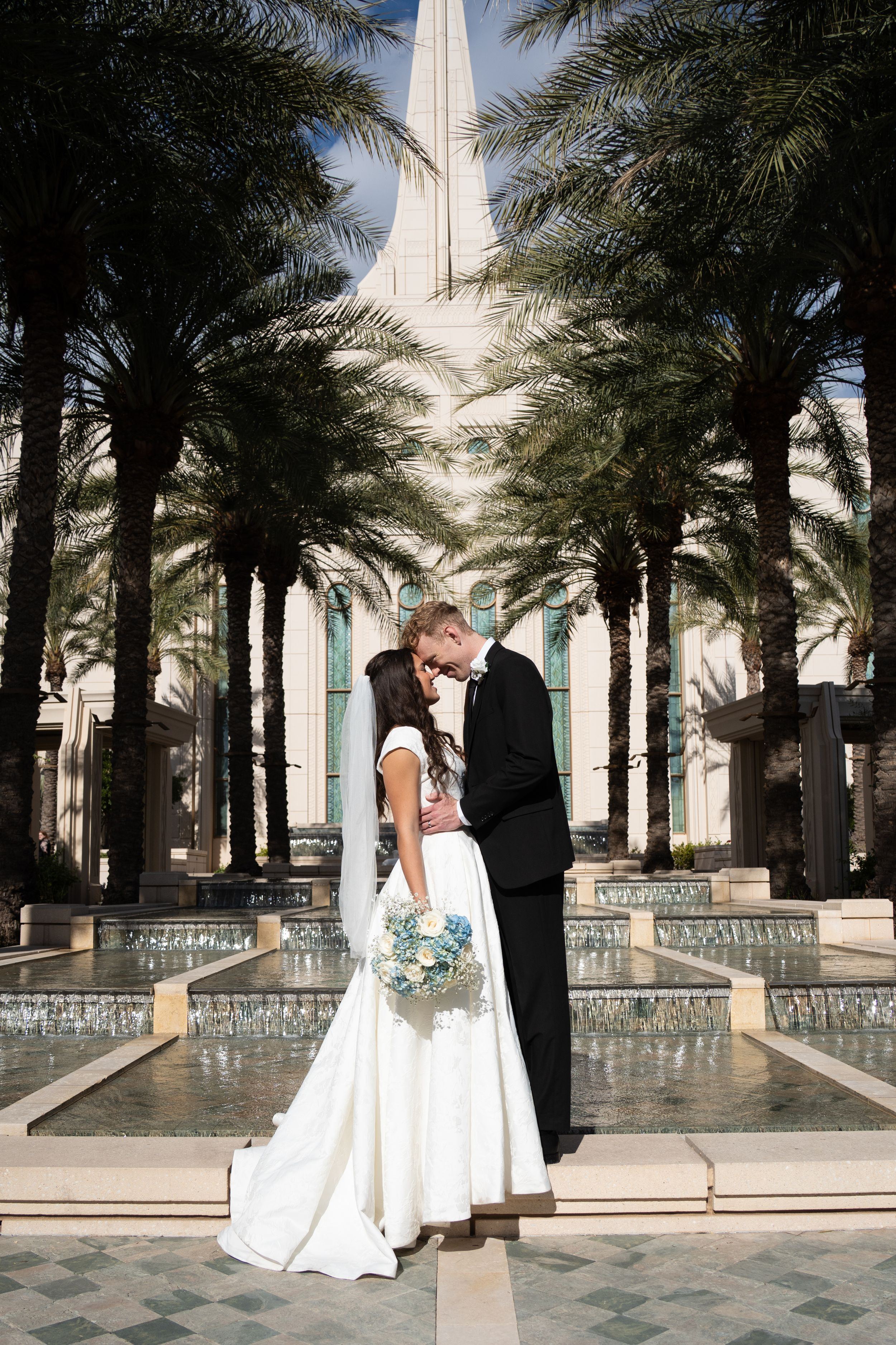 Bride and groom standing close together beneath a grand spired building and rows of palm trees, framed by cascading fountains