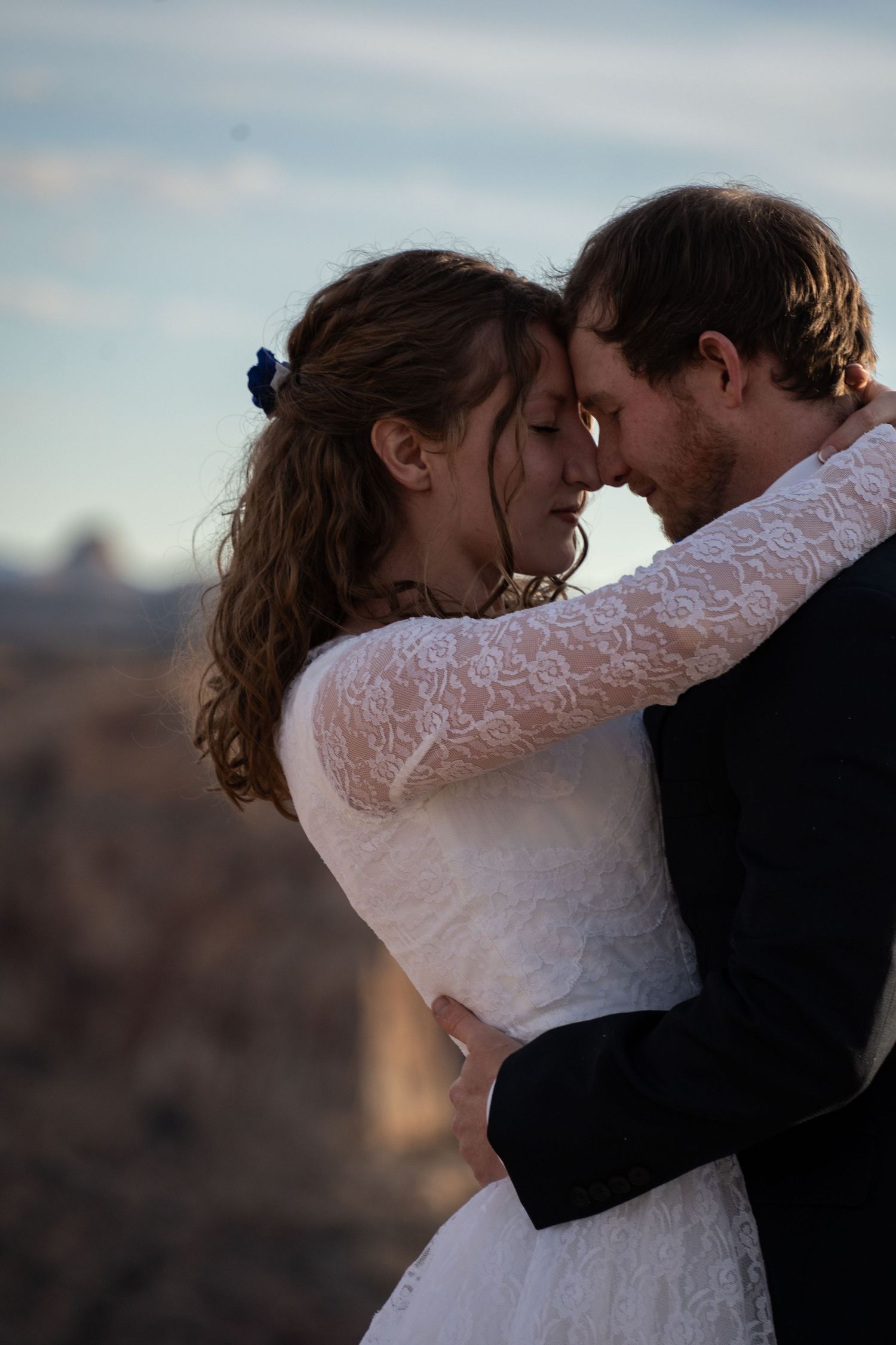 A bride and groom in wedding dress and suit share a romantic forehead touch embrace outdoors with rough canyon backdrop.