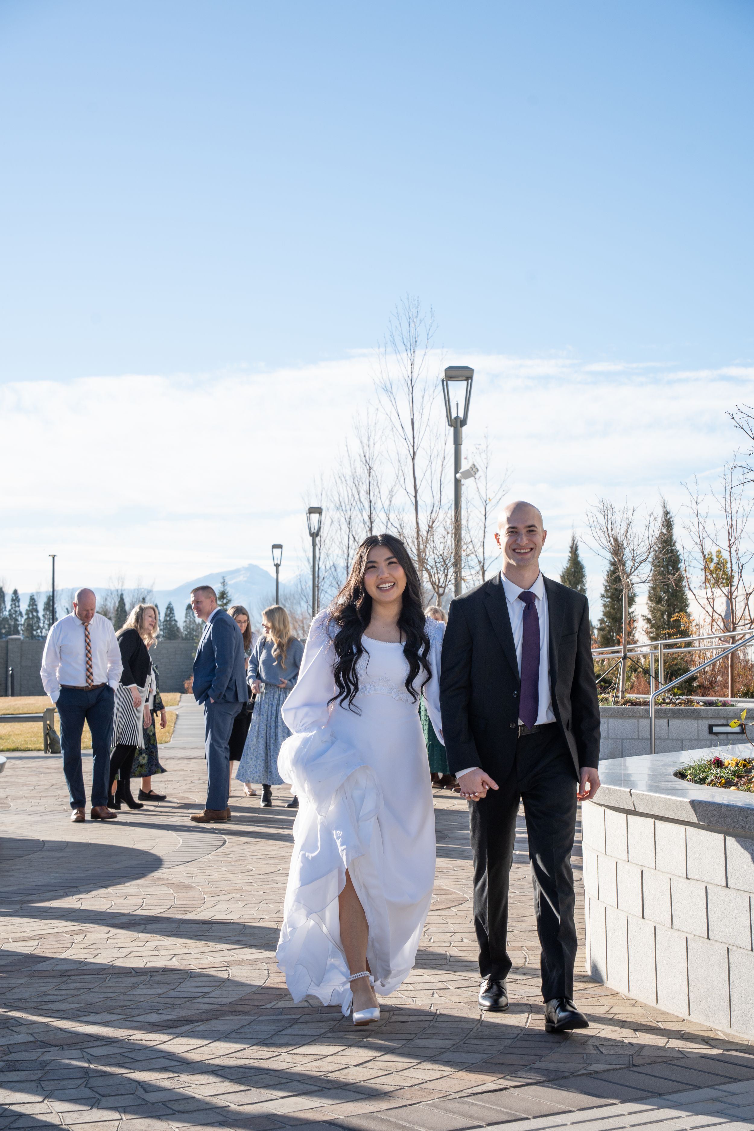 Laughing bride and groom walk around Orem Utah Temple surrounded by family and friends, a Utah temple wedding day from Orem, not Ogden