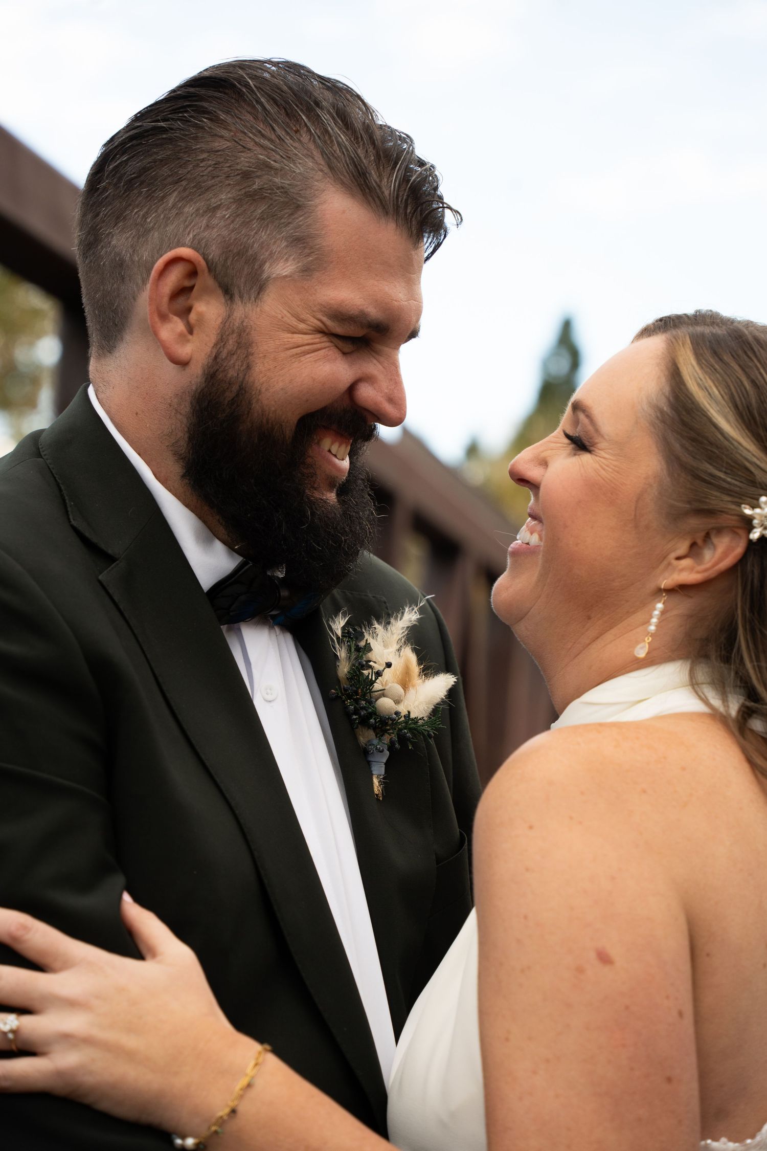 A smiling bride and groom in formal wedding attire share a joyful moment, with the groom wearing a tuxedo and floral boutonniere.